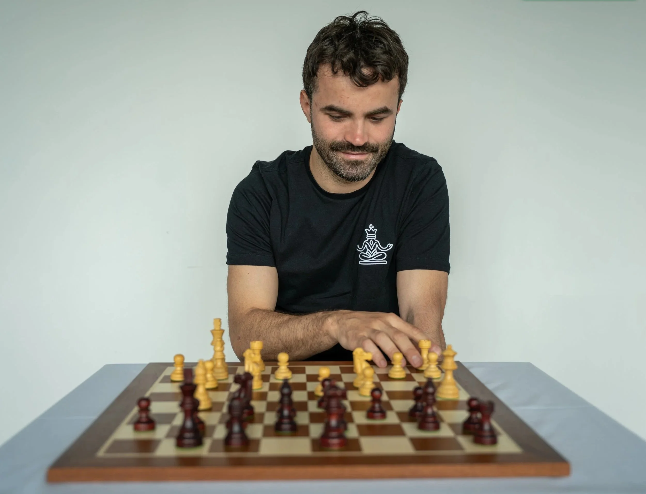 A man with curly dark hair and a beard playing chess on a wooden board while seated at a desk against a plain off-white wall.