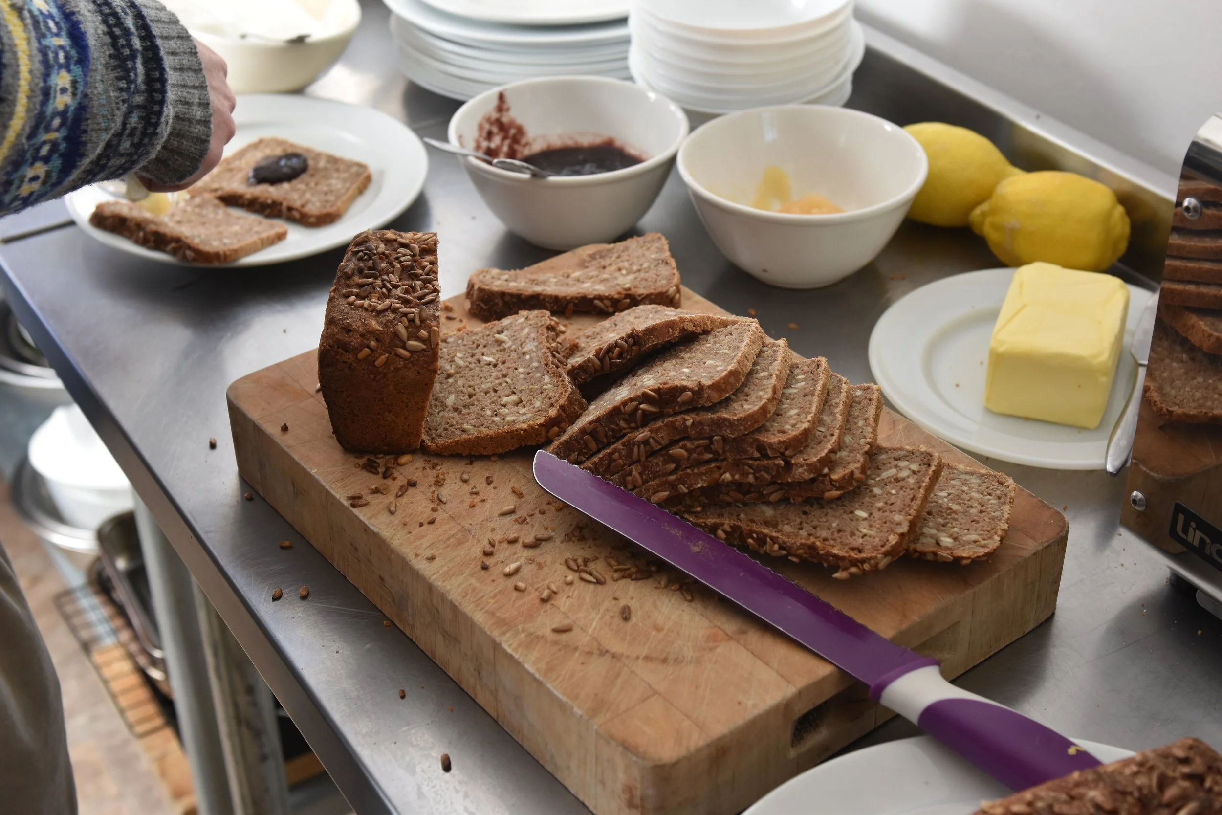 Sliced loaf of multigrain bread, a piece being spread with butter, on a wooden cutting board. In the background, there are bowls of jam, lemon, and butter, with stacked white plates.