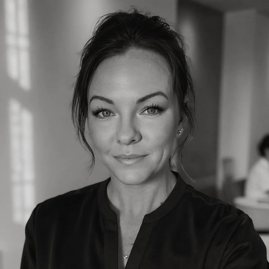 A woman with dark hair, styled in a loose updo, taking a selfie in black and white. She has light-colored eyes, well-defined eyebrows, and is wearing a dark top with a subtle smile.