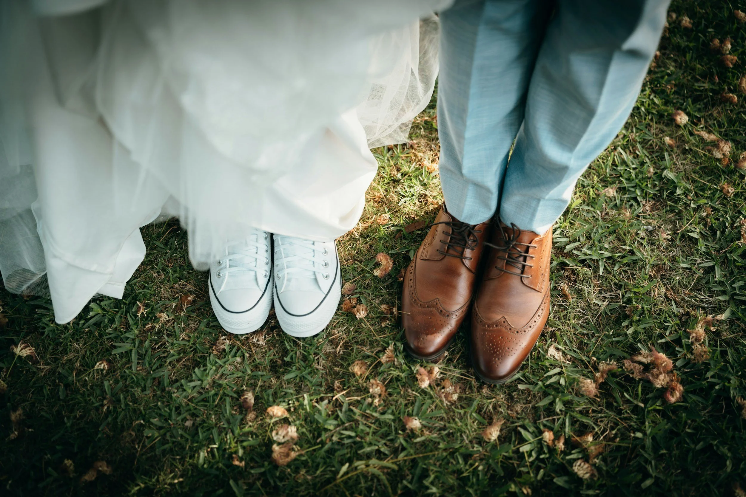 Close-up of a couple's feet standing on grass, showing one person in white sneakers and the other in brown leather dress shoes, with a white dress and blue pants visible.