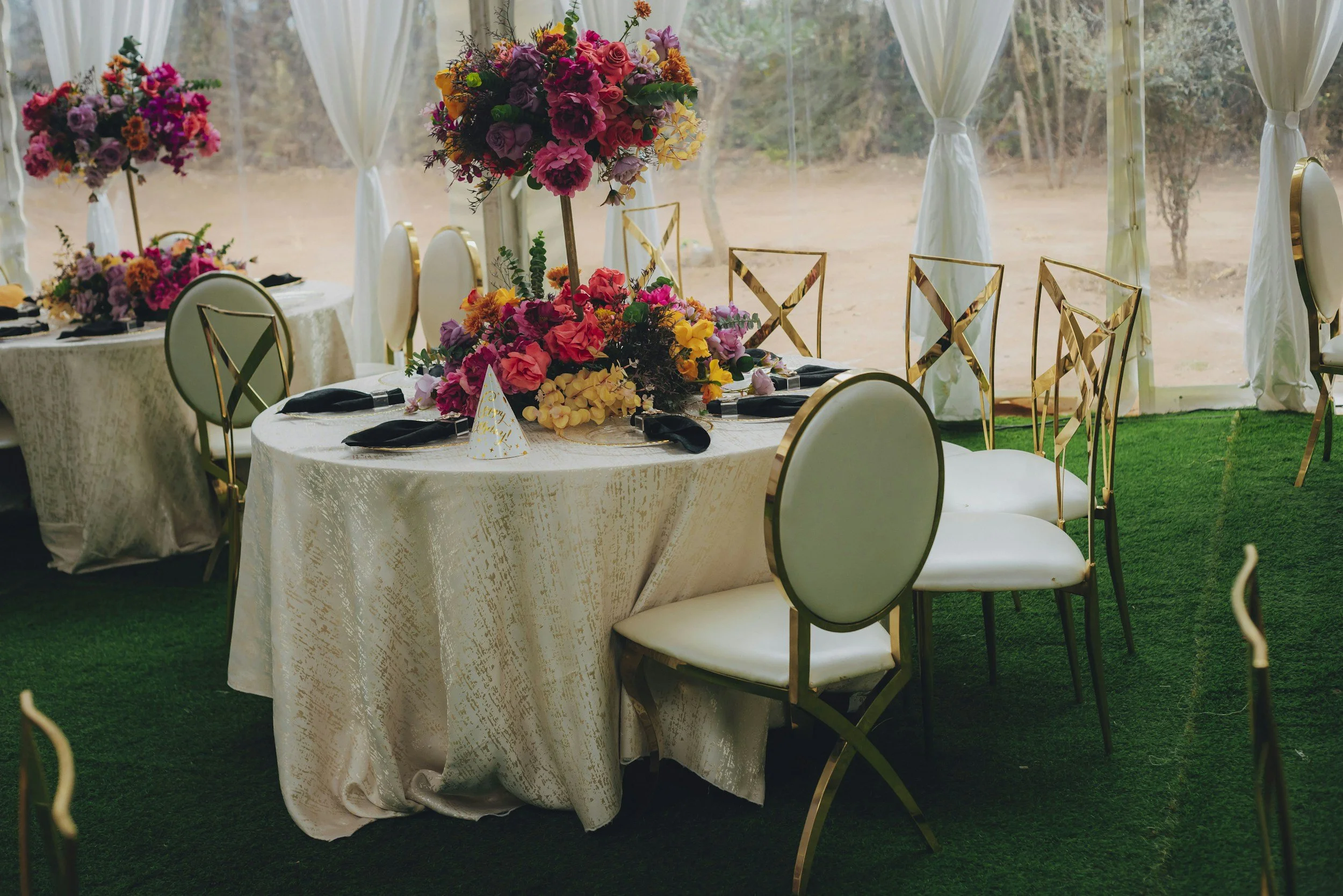 Outdoor wedding ceremony setup with a floral arch in front of a wooden fence, white chairs, and lantern decorations on a grassy lawn.