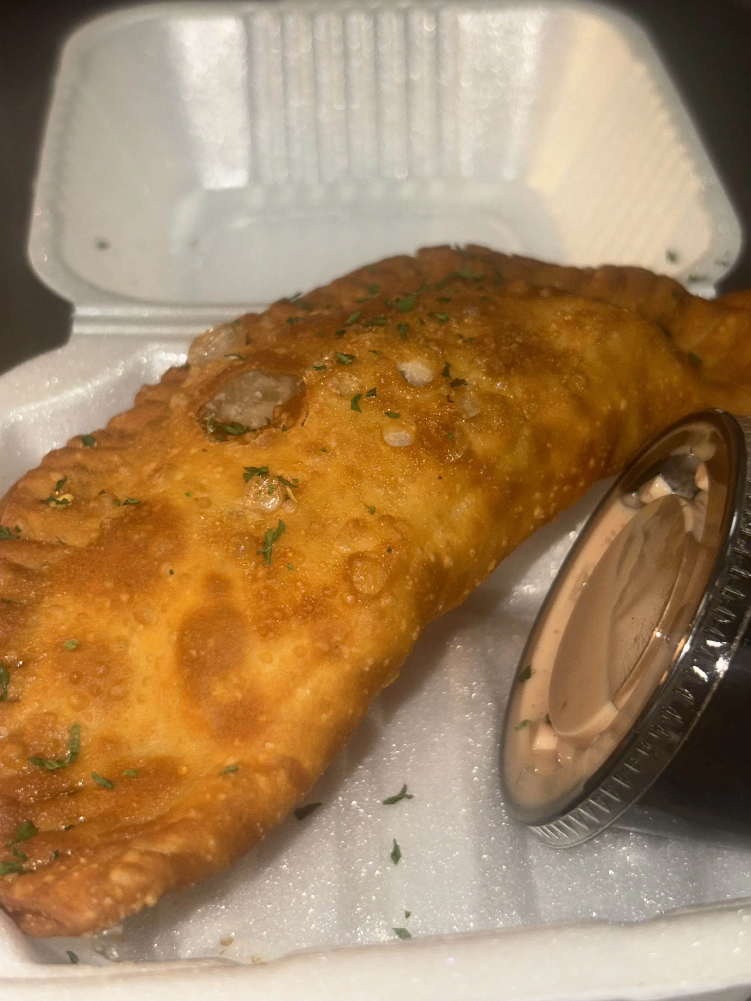 Close-up of a fried fish fillet inside a white foam takeout container, garnished with chopped herbs, with a small container of tartar sauce beside it.