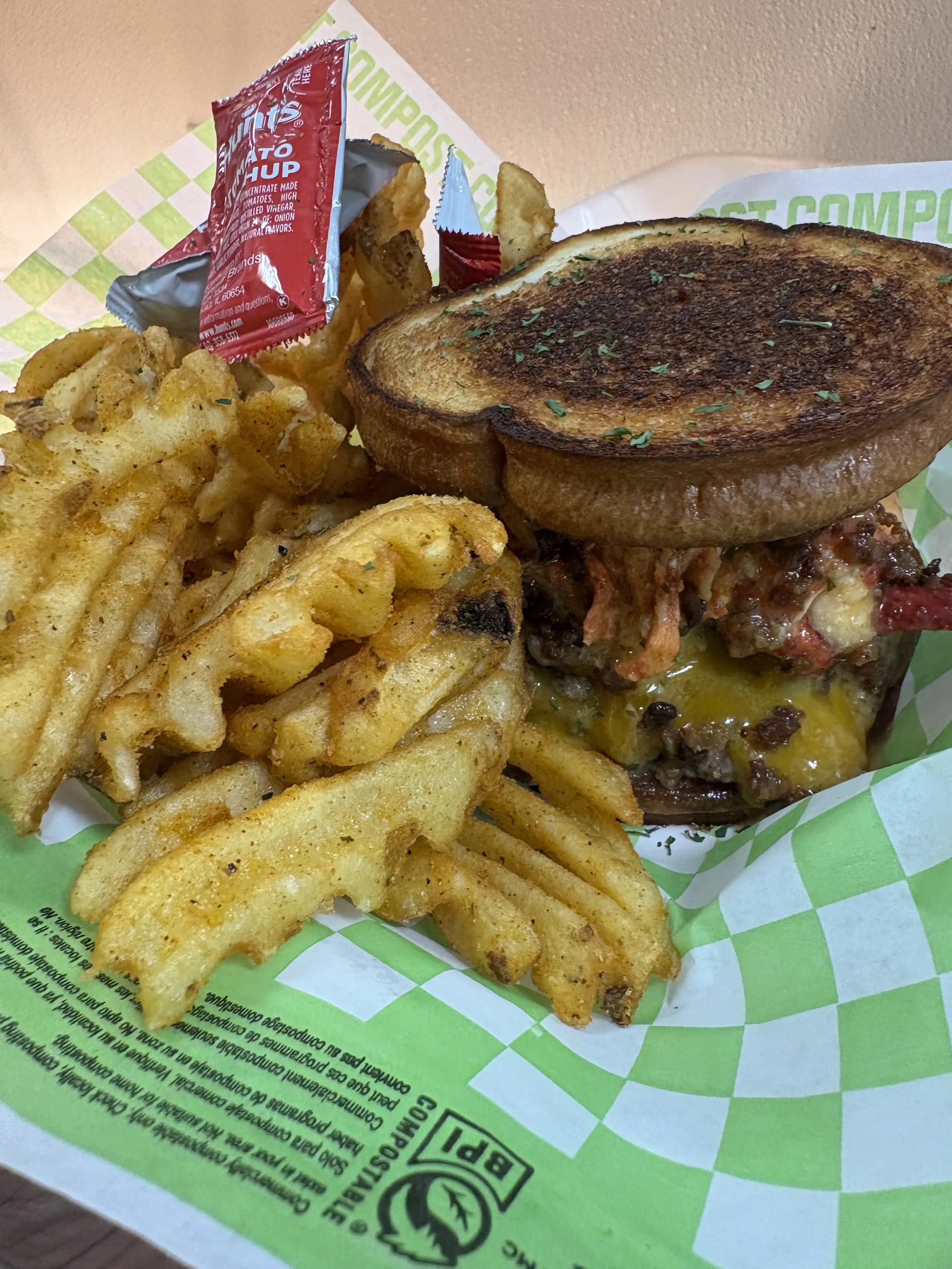 Close-up photo of a breakfast sandwich with a toasted bun, bacon, and melted cheese, served with crinkle-cut French fries and a packet of ketchup in a green and white checkered paper tray.