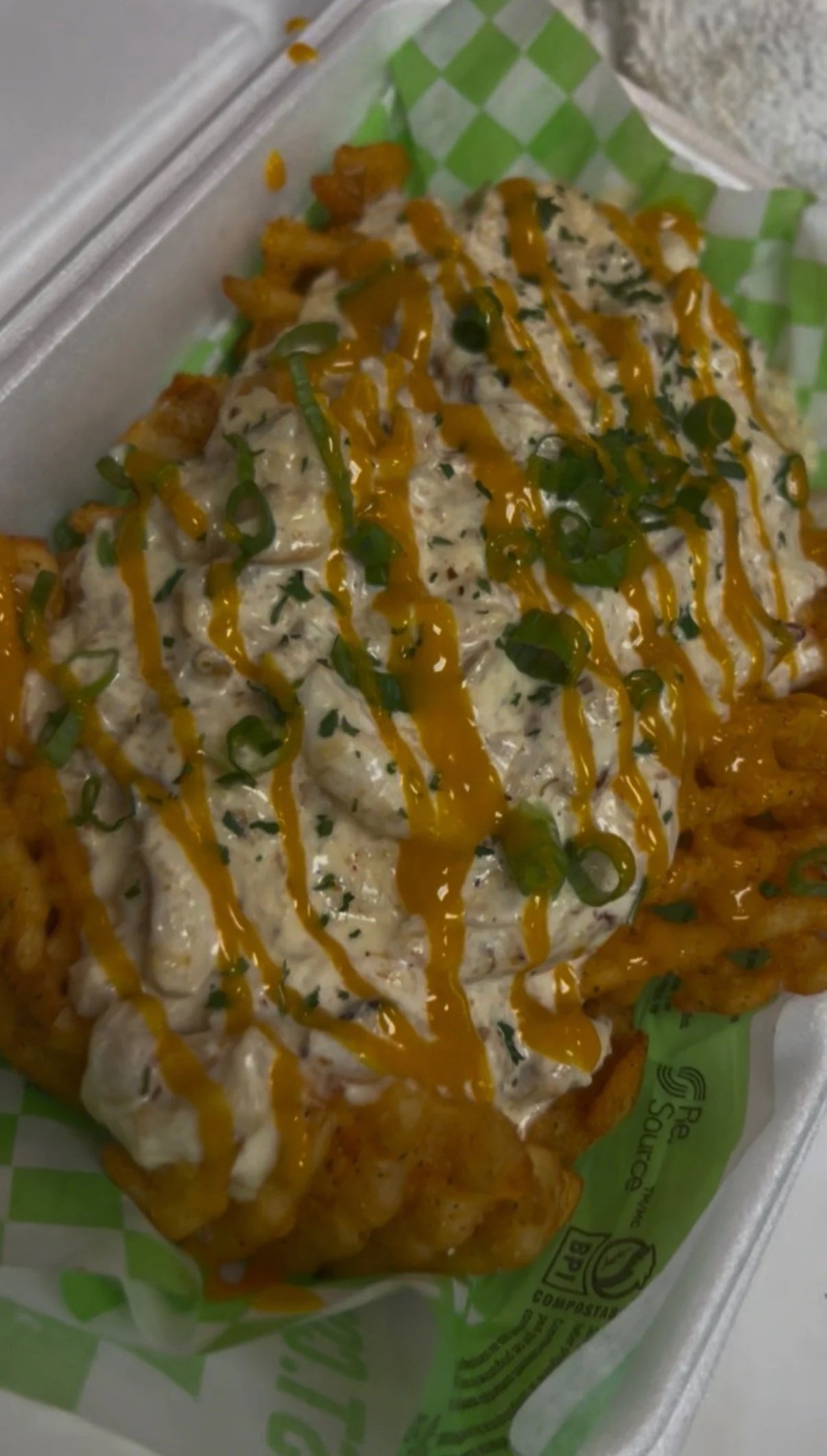Fried food topped with a creamy sauce, chopped green onions, and orange sauce, placed on green and white wrapped paper inside a paper food tray.