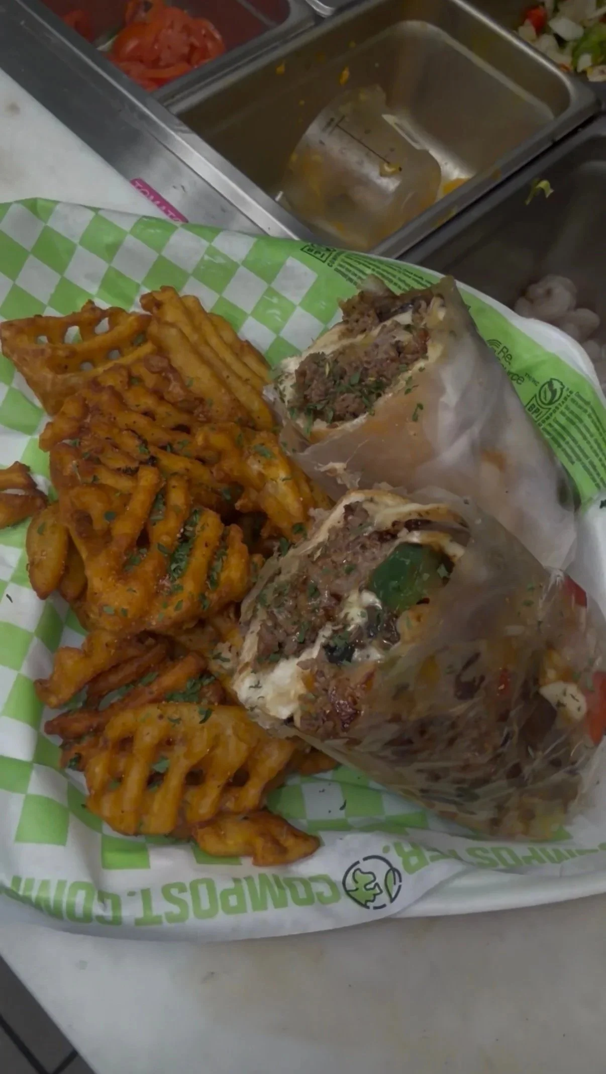 Waffle fries, two half-foiled wrapped beef sandwiches, and chopped vegetables on a green and white checkered paper in a fast-food restaurant. In the background, there is a metal food counter with kitchen ingredients.