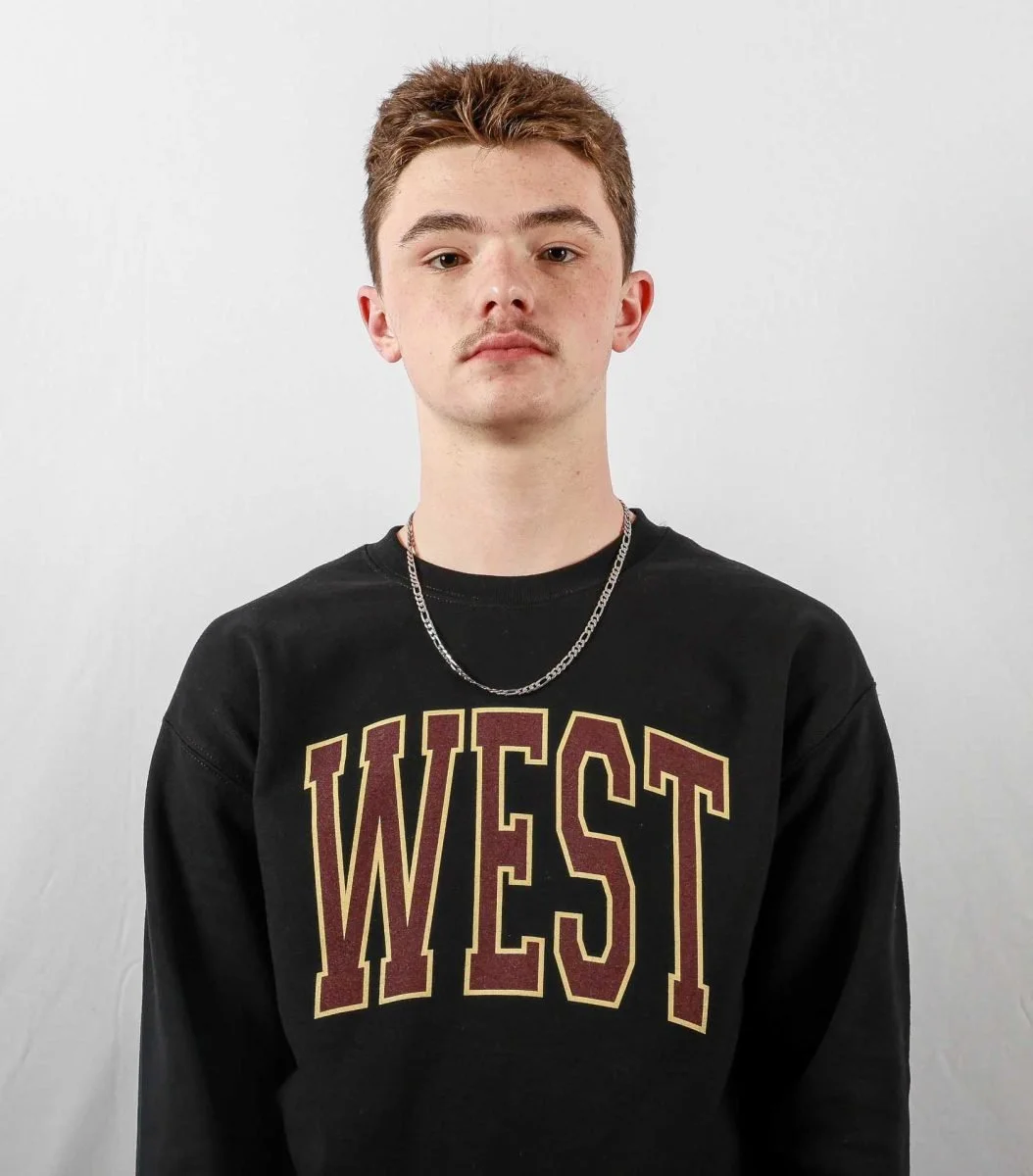 A young man with short, light brown hair, light skin, and a slight mustache, wearing a black sweatshirt with 'WEST' printed in large maroon and gold letters on the front, standing against a plain white background.