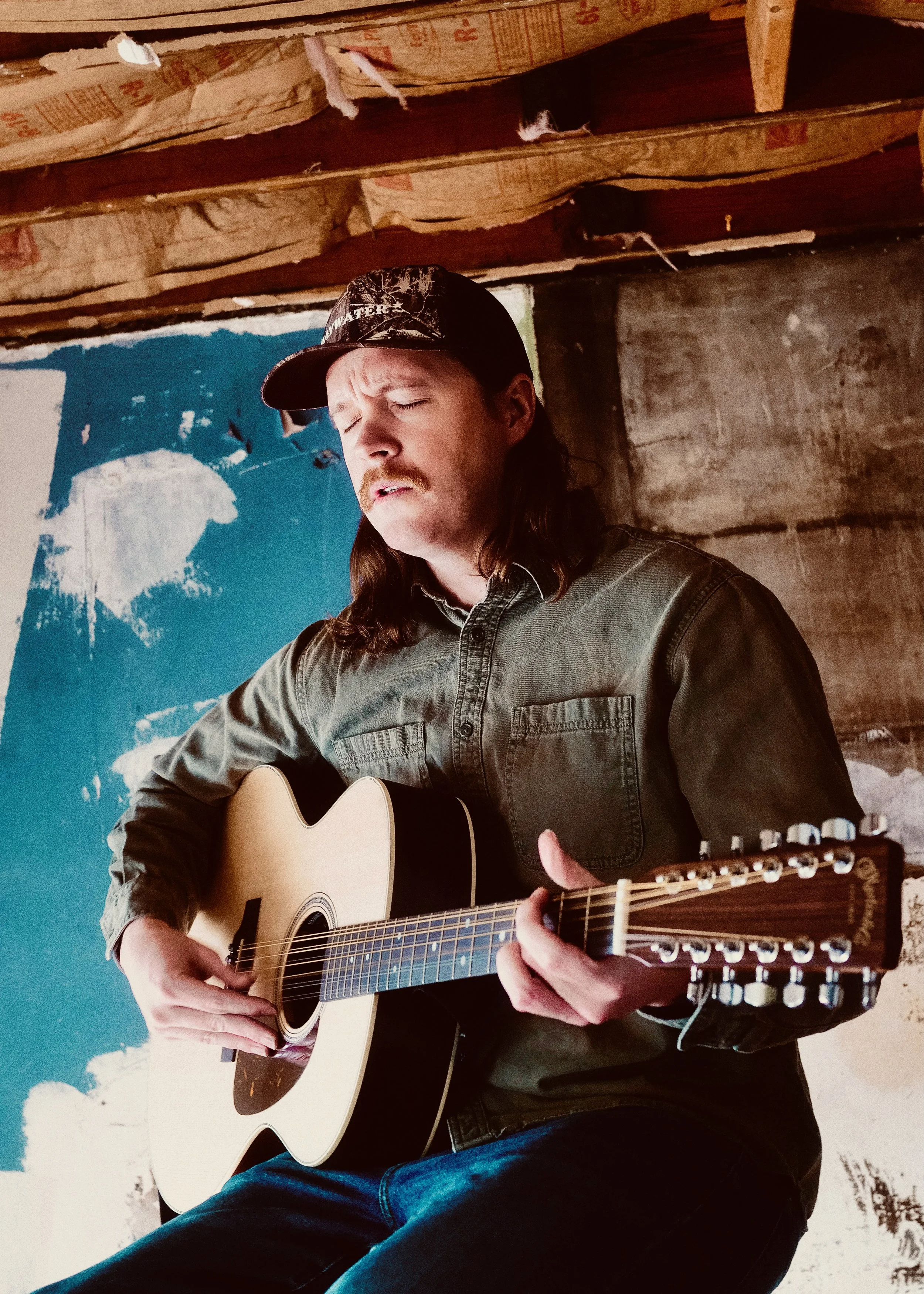 Clayton Nile Young with closed eyes, playing an acoustic guitar. He is wearing a camouflage cap and a gray button-up shirt, standing inside a rustic space with a partially painted blue wall and exposed wooden beams.