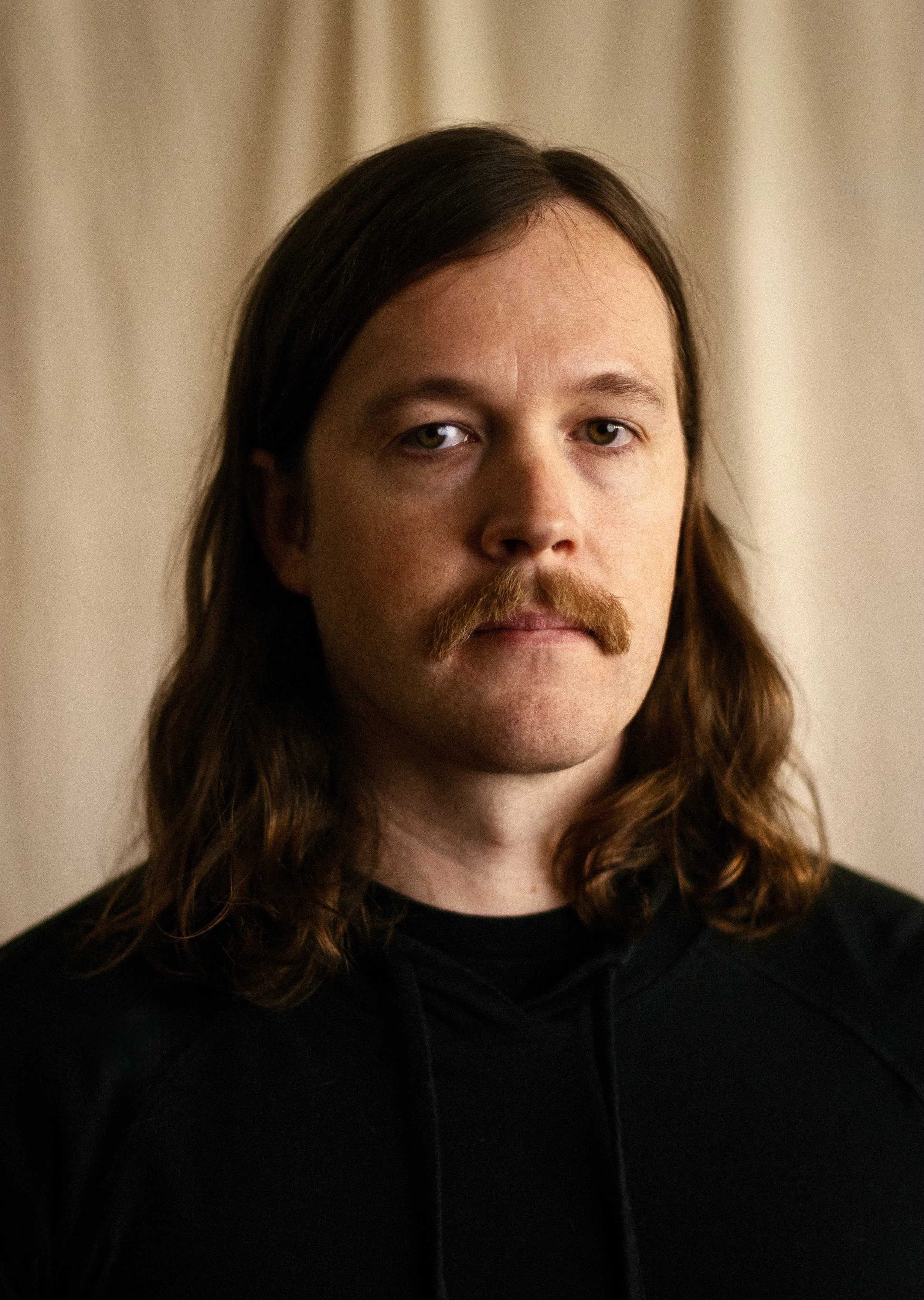 Clayton Nile Young with long brown hair, a mustache, and a goatee, wearing a black shirt, standing in front of a neutral background.