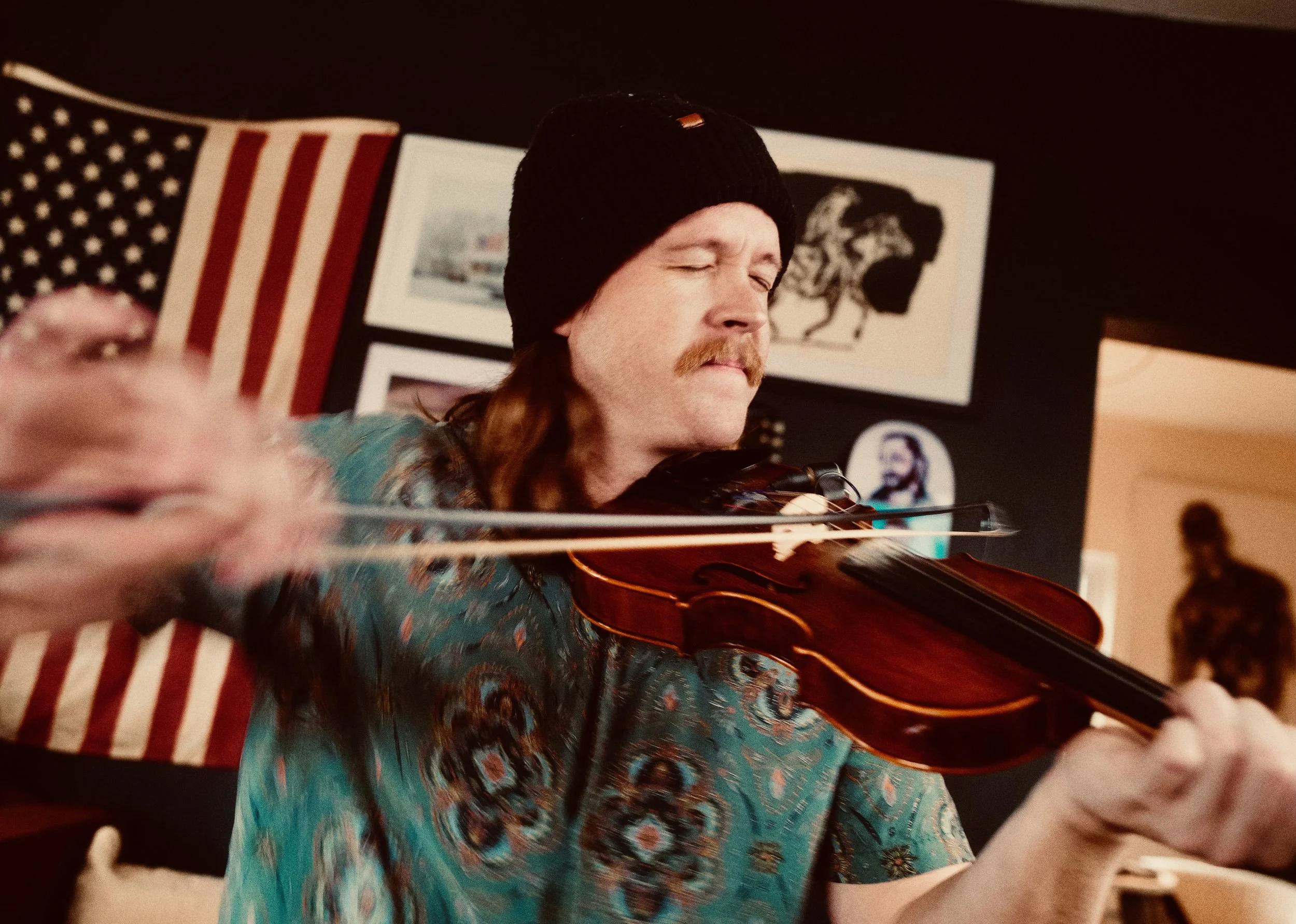Clayton Nile Young with long reddish-brown hair, a mustache, wearing a black beanie, playing a fiddle, with American flags and artwork in the background.