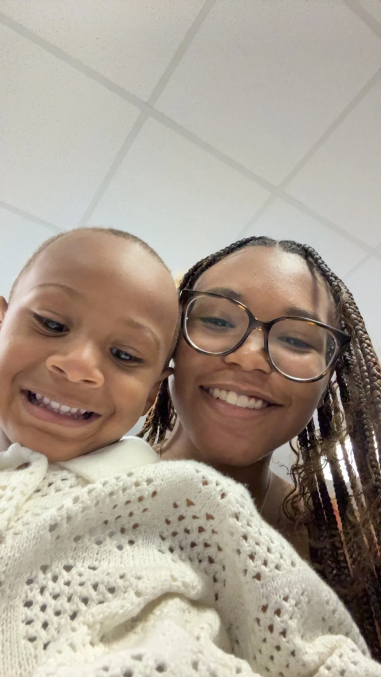 A smiling woman with glasses and braided hair taking a selfie with a young boy with a buzz cut, both appearing happy, against a ceiling with ceiling tiles.