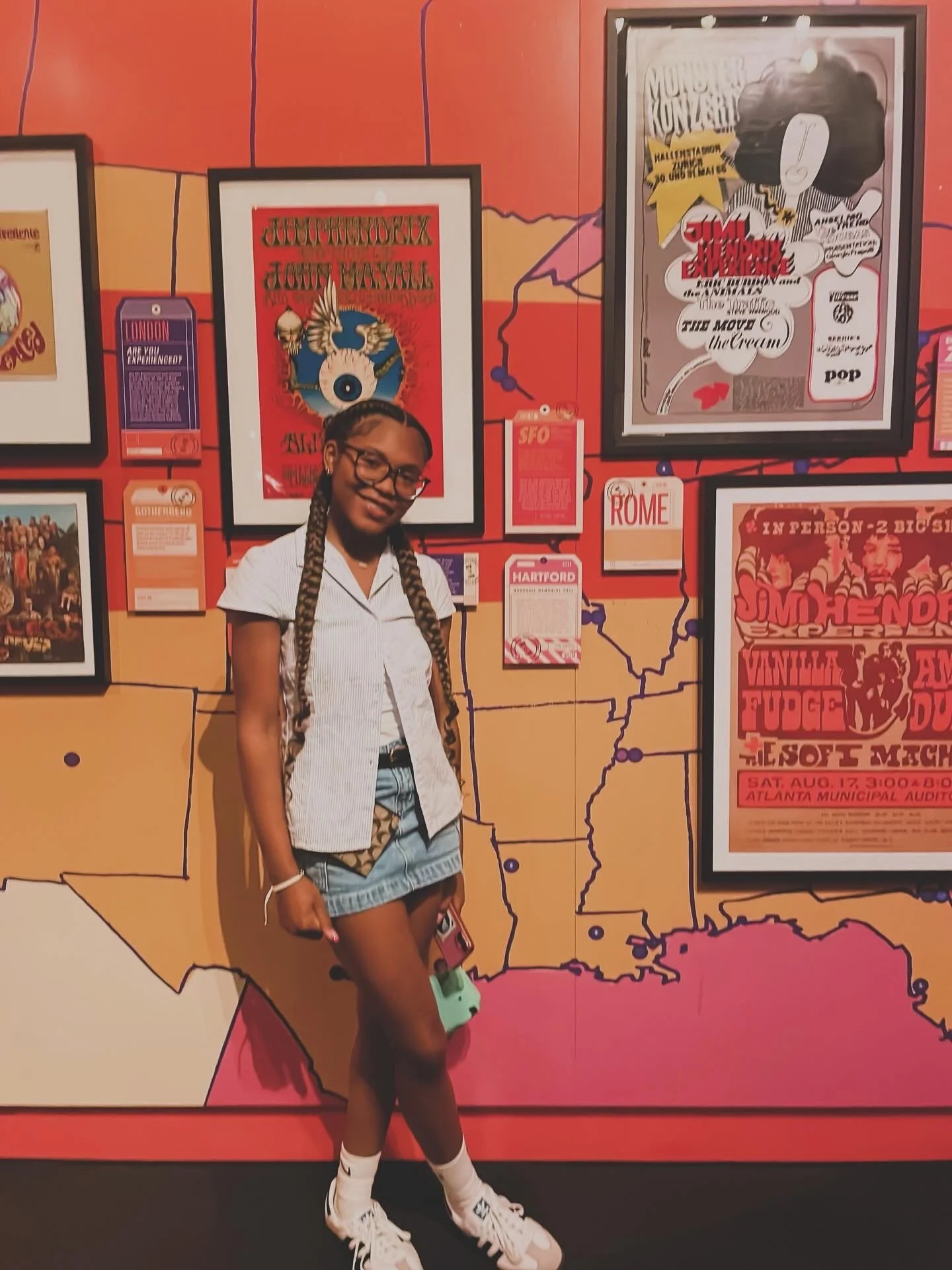 Young woman with braided hair and glasses standing in front of a colorful mural of the United States map with various posters and event flyers. She is smiling, wearing a striped shirt, denim shorts, white socks, and sneakers, holding a pink phone and a small green bag.