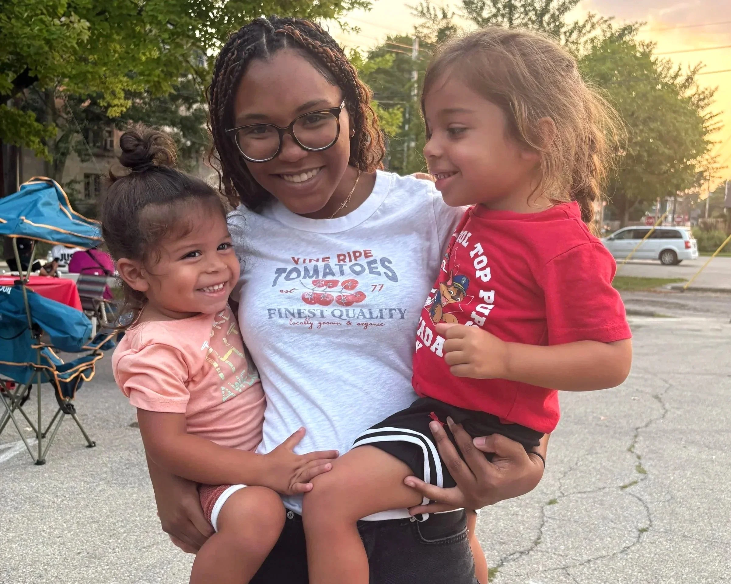 Three smiling children being held by a smiling woman outdoors during sunset, with trees and parked cars in the background.