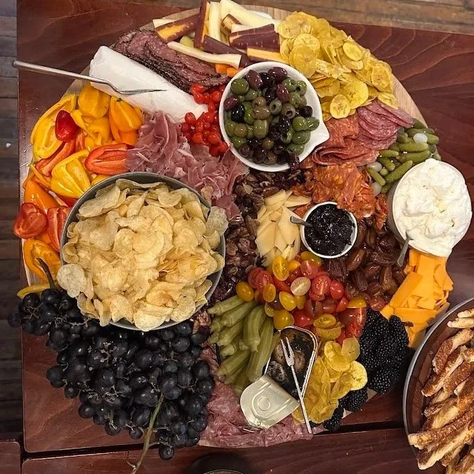 A circular charcuterie board with various cheeses, meats, crackers, vegetables, fruits, and toppings, arranged in sections for a snack platter.