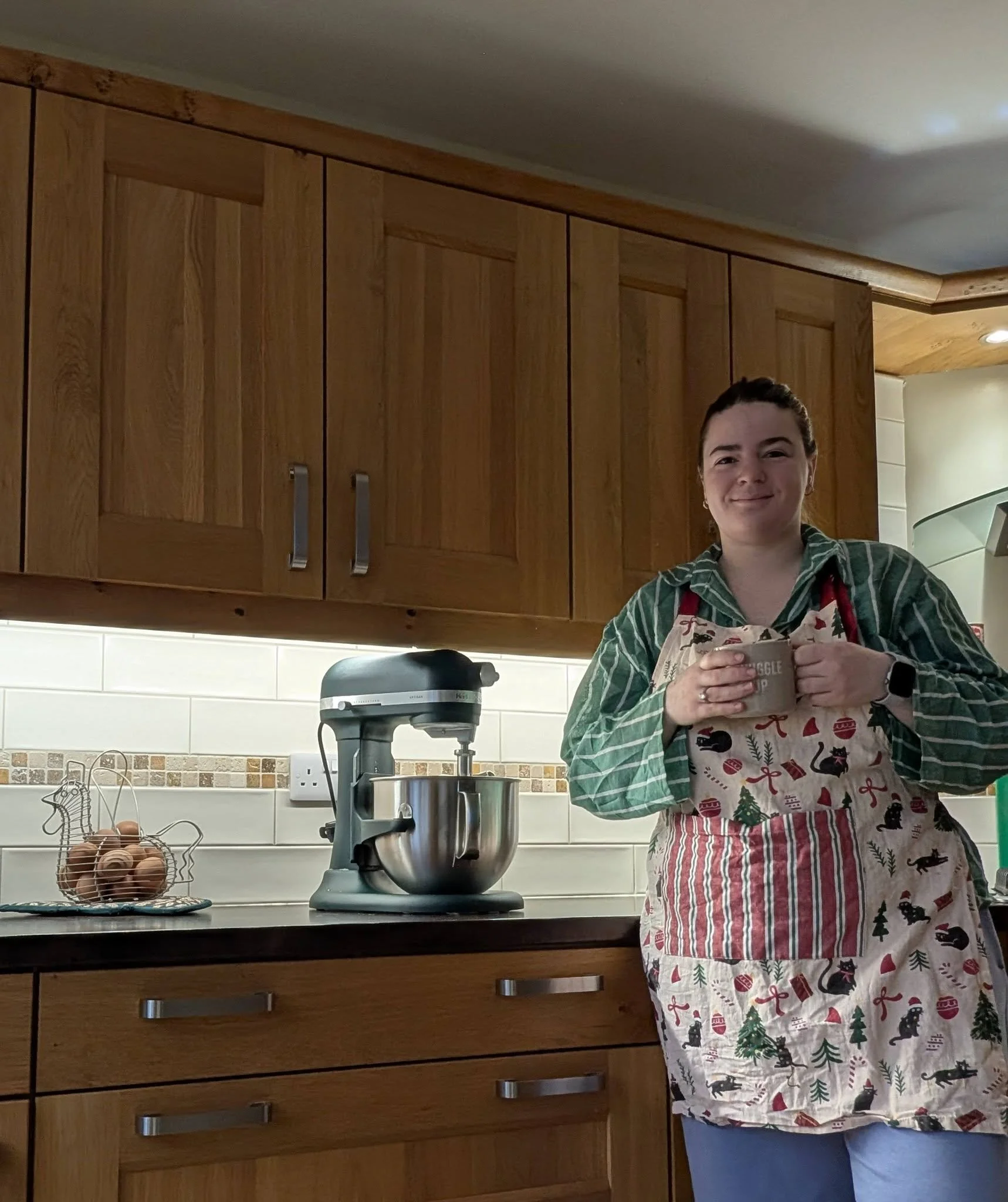 A woman wearing a Christmas-themed apron stands in a kitchen, smiling and holding a mug. The kitchen features wooden cabinets, a stand mixer on the counter, and a basket of eggs.