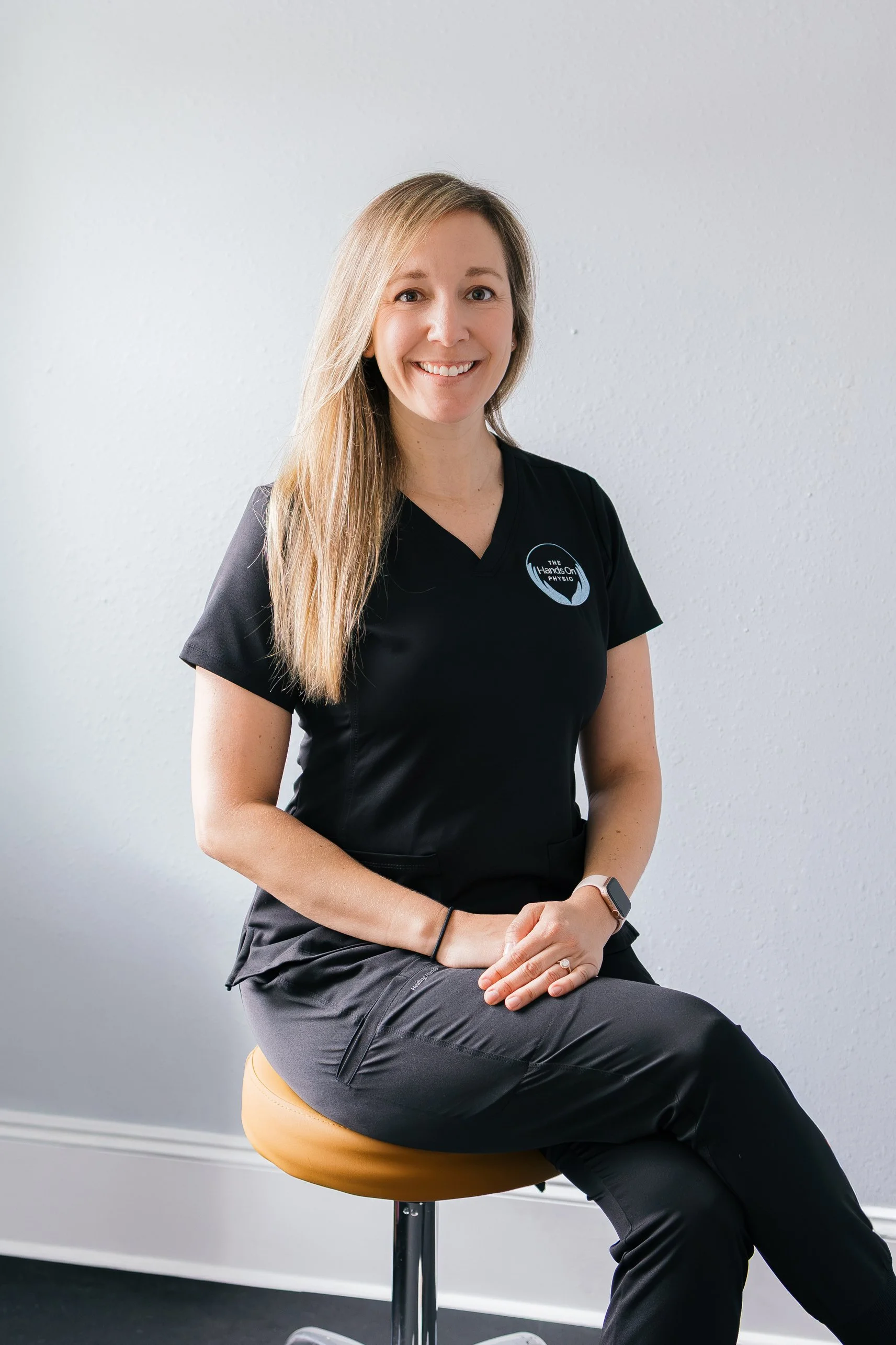 A woman with long blonde hair sitting on a beige round stool, smiling, wearing gray and black athletic clothing, and a smartwatch, in front of a plain white wall.