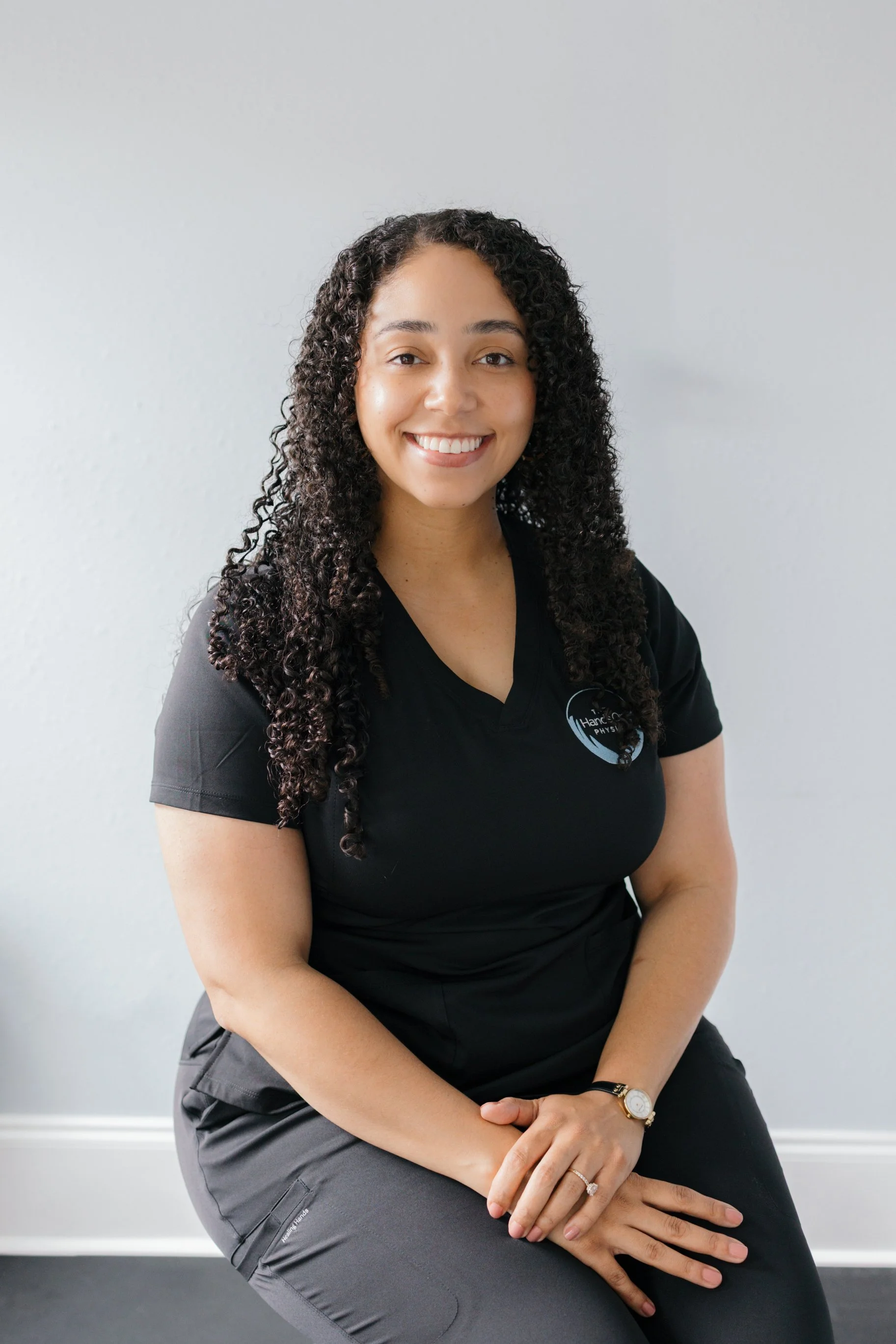 A woman with curly hair and a bright smile sitting against a plain white wall, wearing a black shirt with a logo on it, gray pants, and a watch.