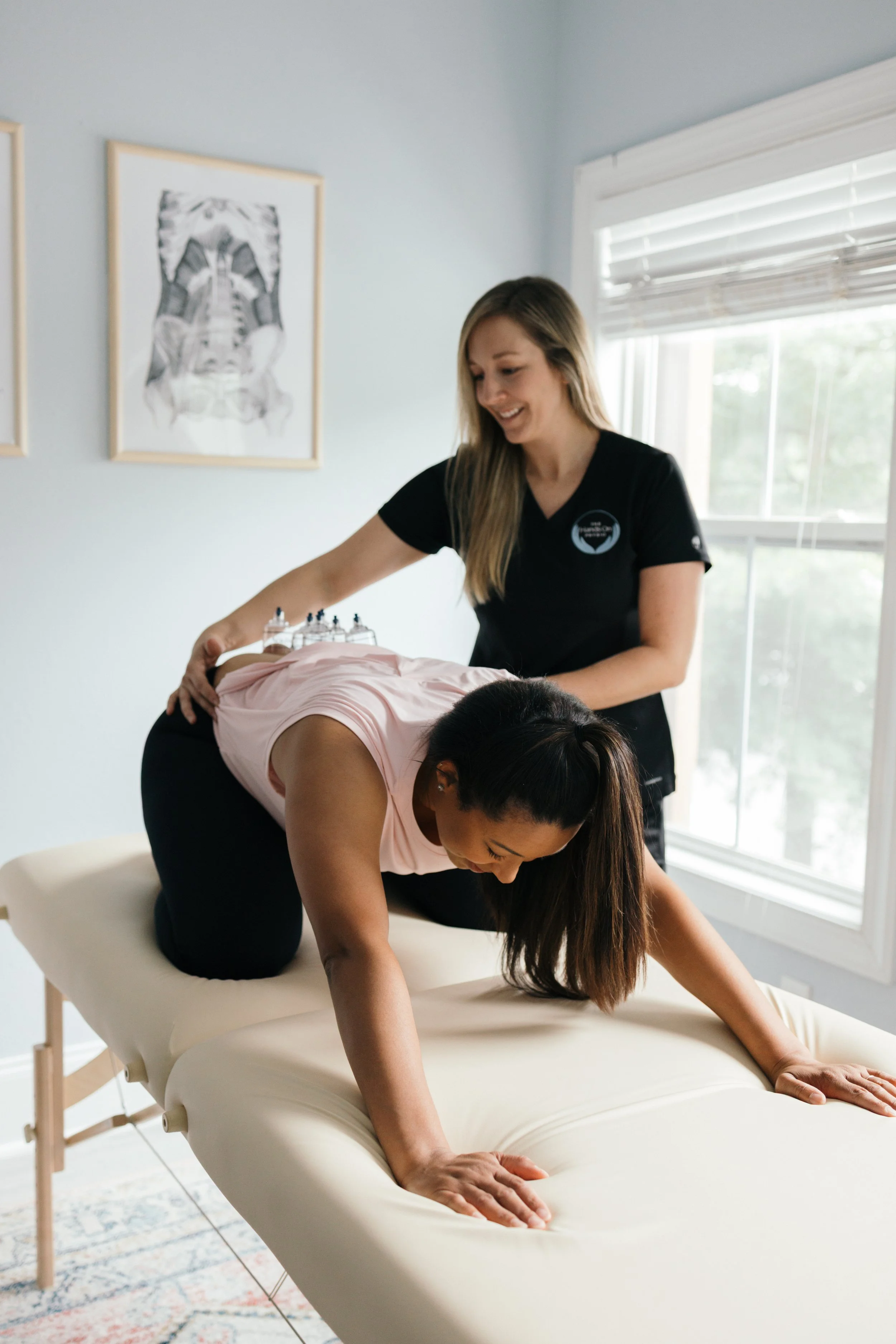 A woman in a pink shirt and black pants is on her hands and knees on a massage table, receiving a back massage from a therapist. The therapist, with long hair and a black shirt, is smiling. The room has natural light from a window and artwork on the wall.