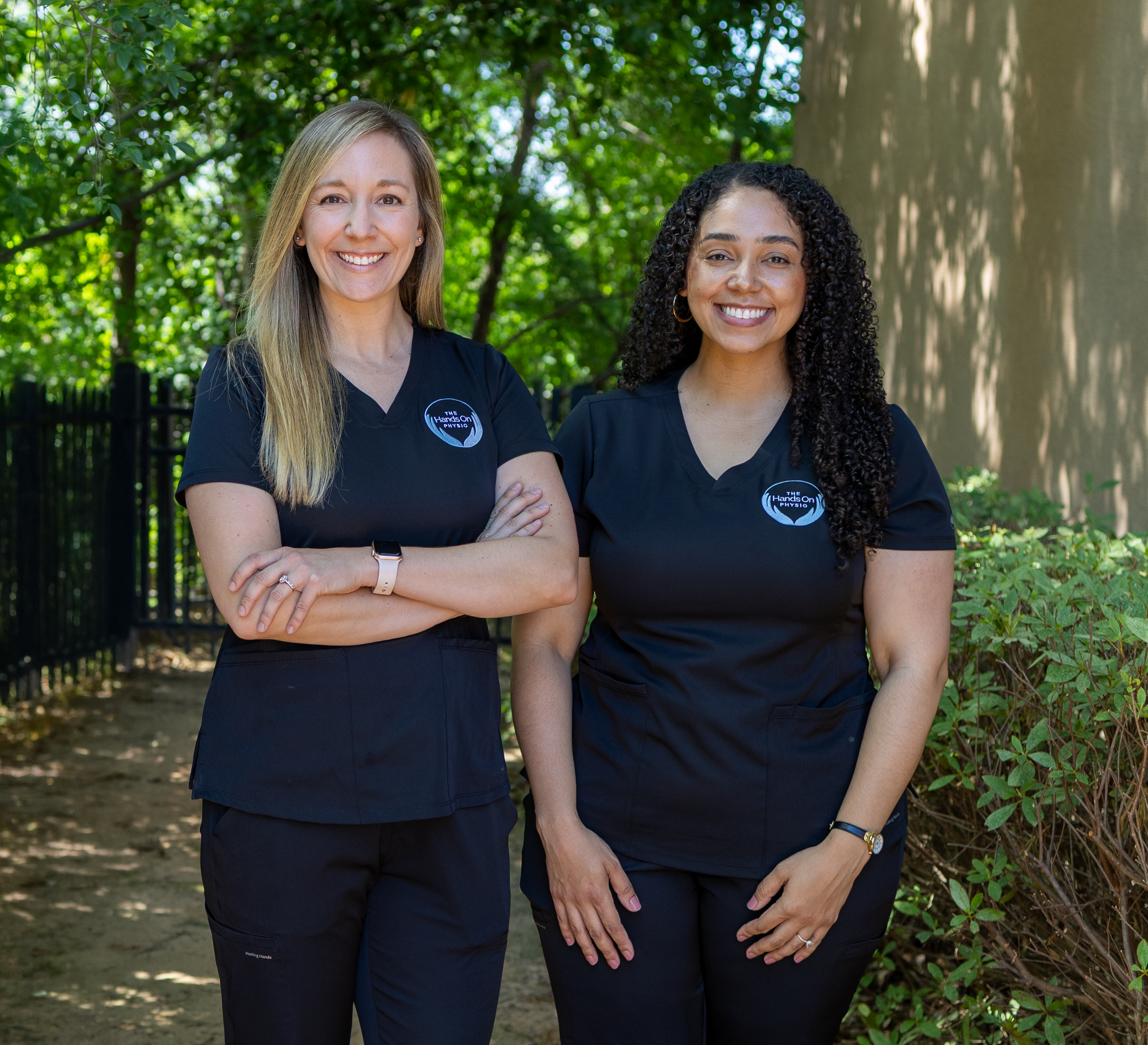 Two women in black medical scrubs standing outdoors among green trees, smiling at the camera.
