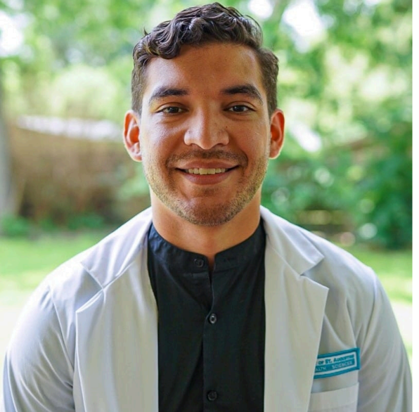 Young male healthcare professional in black scrubs and white lab coat smiling outdoors with green trees in the background.