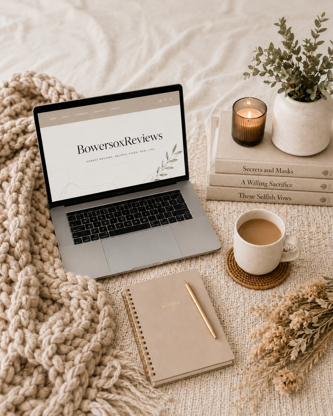 A cozy workspace with a laptop displaying 'BowersoxReviews', a mug of coffee on a woven coaster, a stack of books, a lit candle, a potted plant, a notebook with a pen, a candle, and dried flowers on a soft textured blanket.
