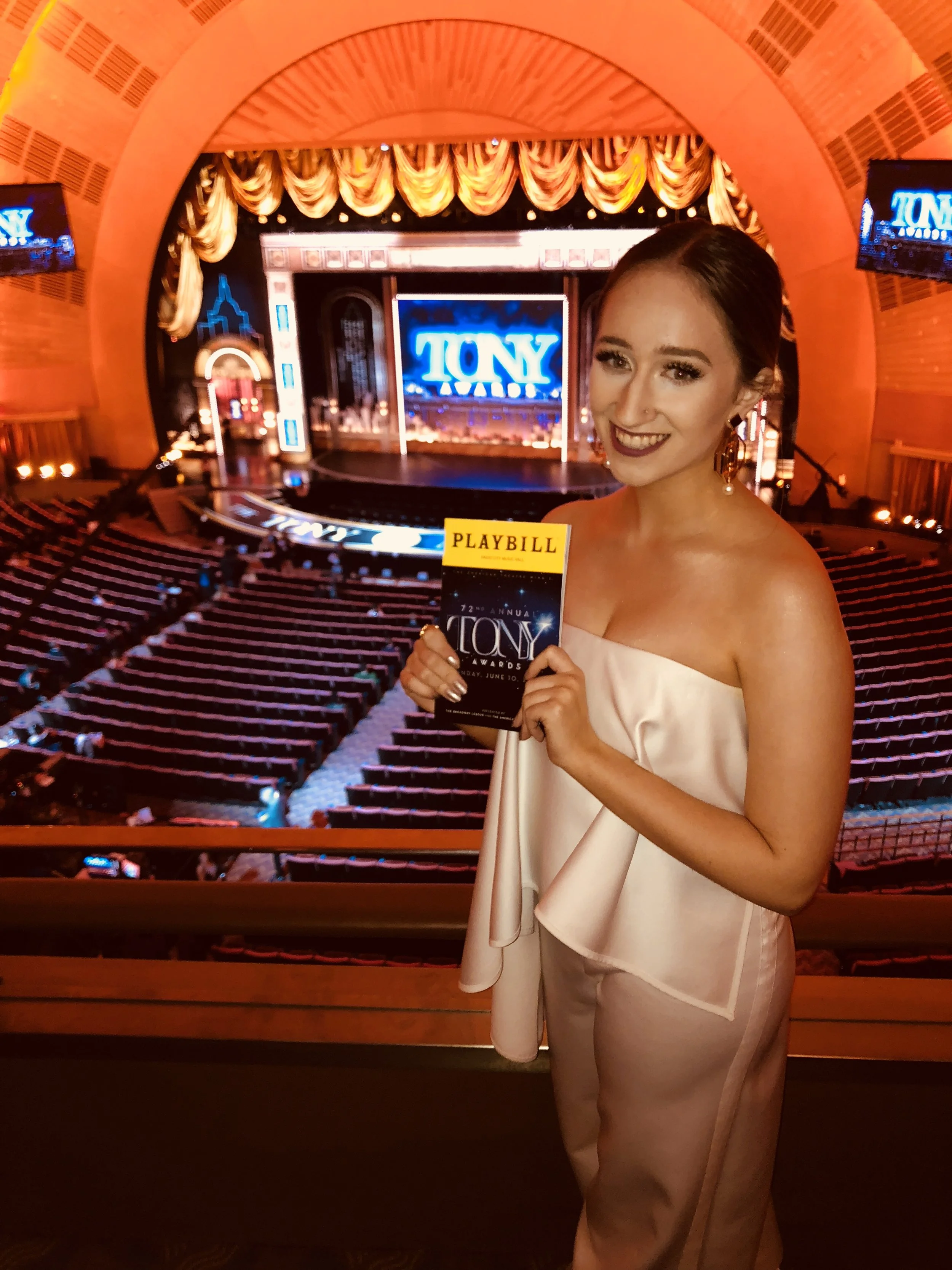 A woman in a light-colored dress holding a Tony Awards playbill, standing in front of a stage with a large illuminated sign that says 'TONY' at an awards show event.