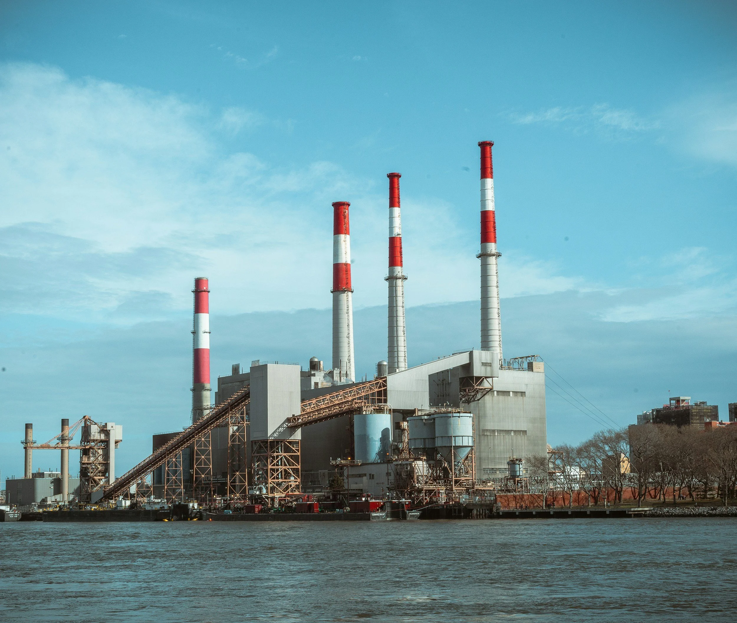 Industrial factory with tall smokestacks painted red and white, located beside a body of water, with a clear blue sky above.