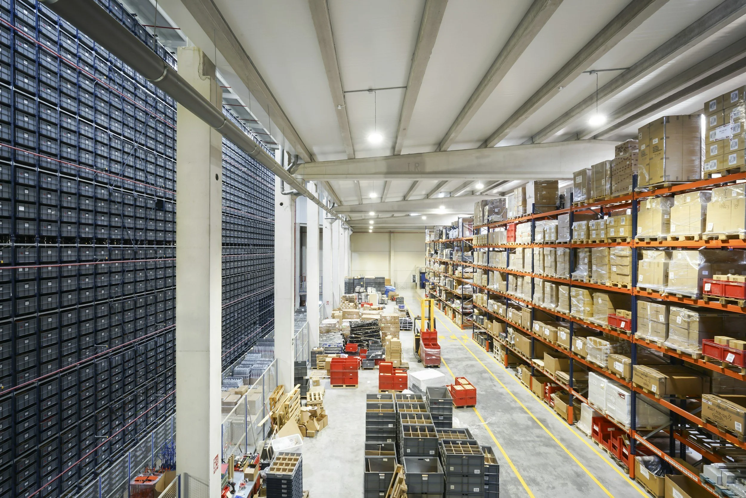 A large warehouse with high shelves filled with boxes and plastic containers. The warehouse has a high ceiling with exposed beams and bright lighting.