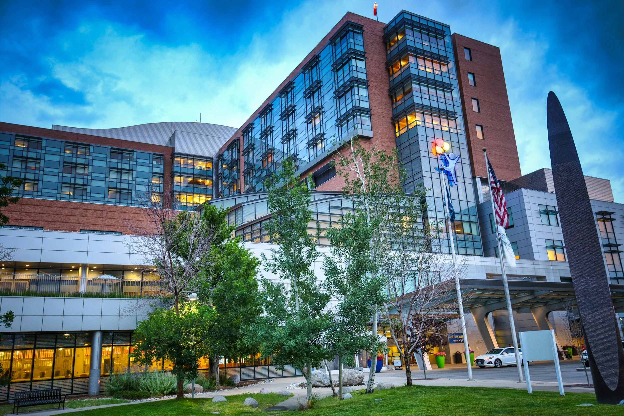Modern multi-story building with glass windows, flags, and a small park area with trees and a bench in the foreground.