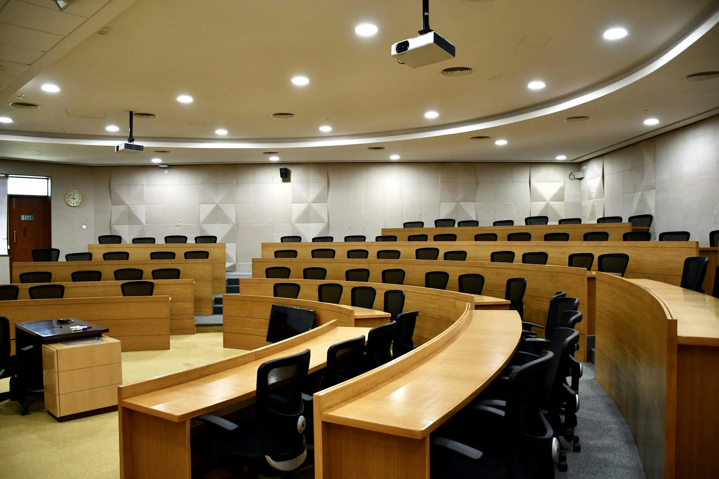 A large conference room with curved wooden tables and black office chairs. The ceiling has multiple recessed lights and two mounted projectors. There's a clock on the wall near a door, and several small computer monitors on the tables.