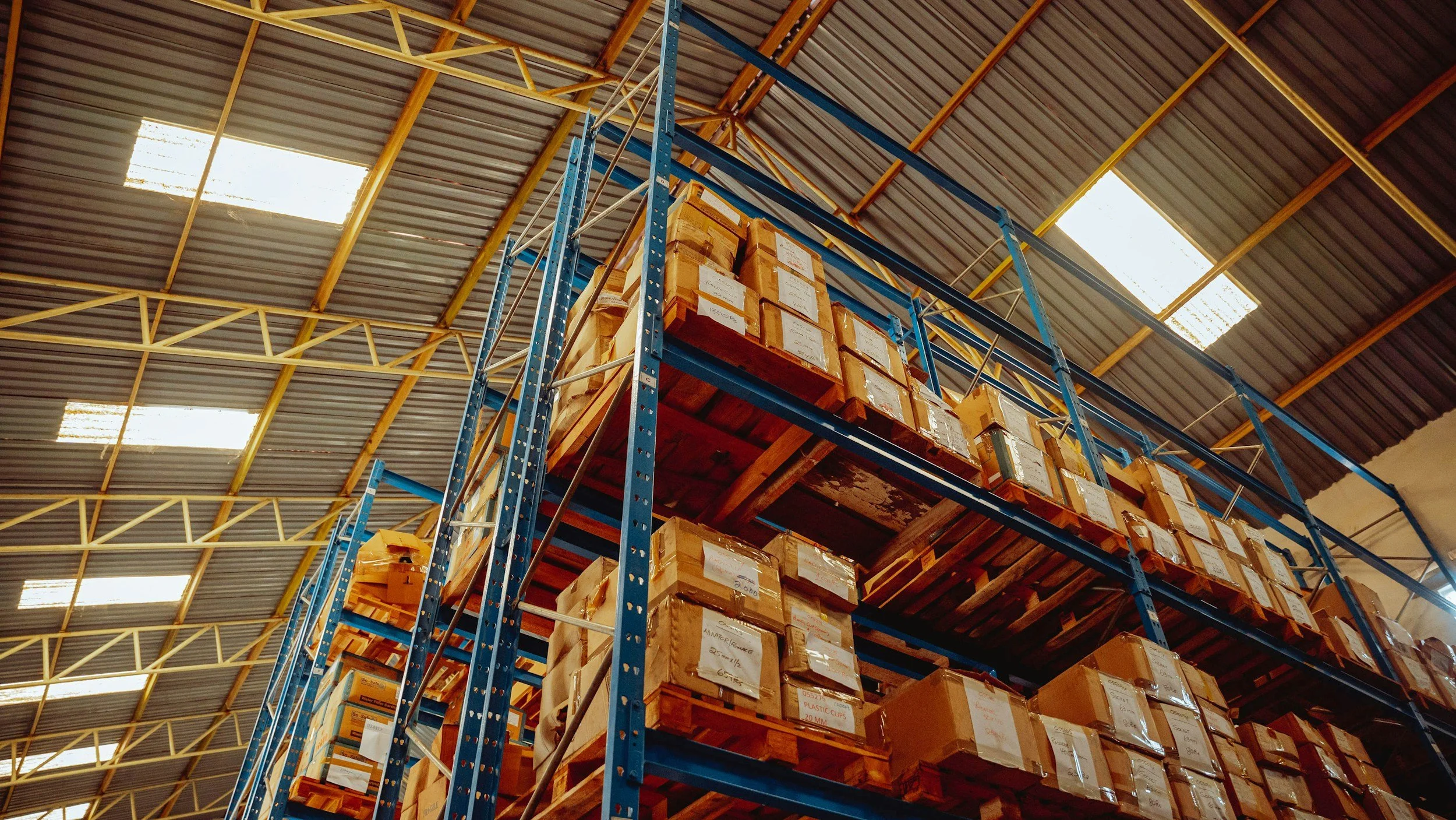View of a warehouse with high blue steel shelves filled with packed boxes. The ceiling is industrial with skylights and metal beams.