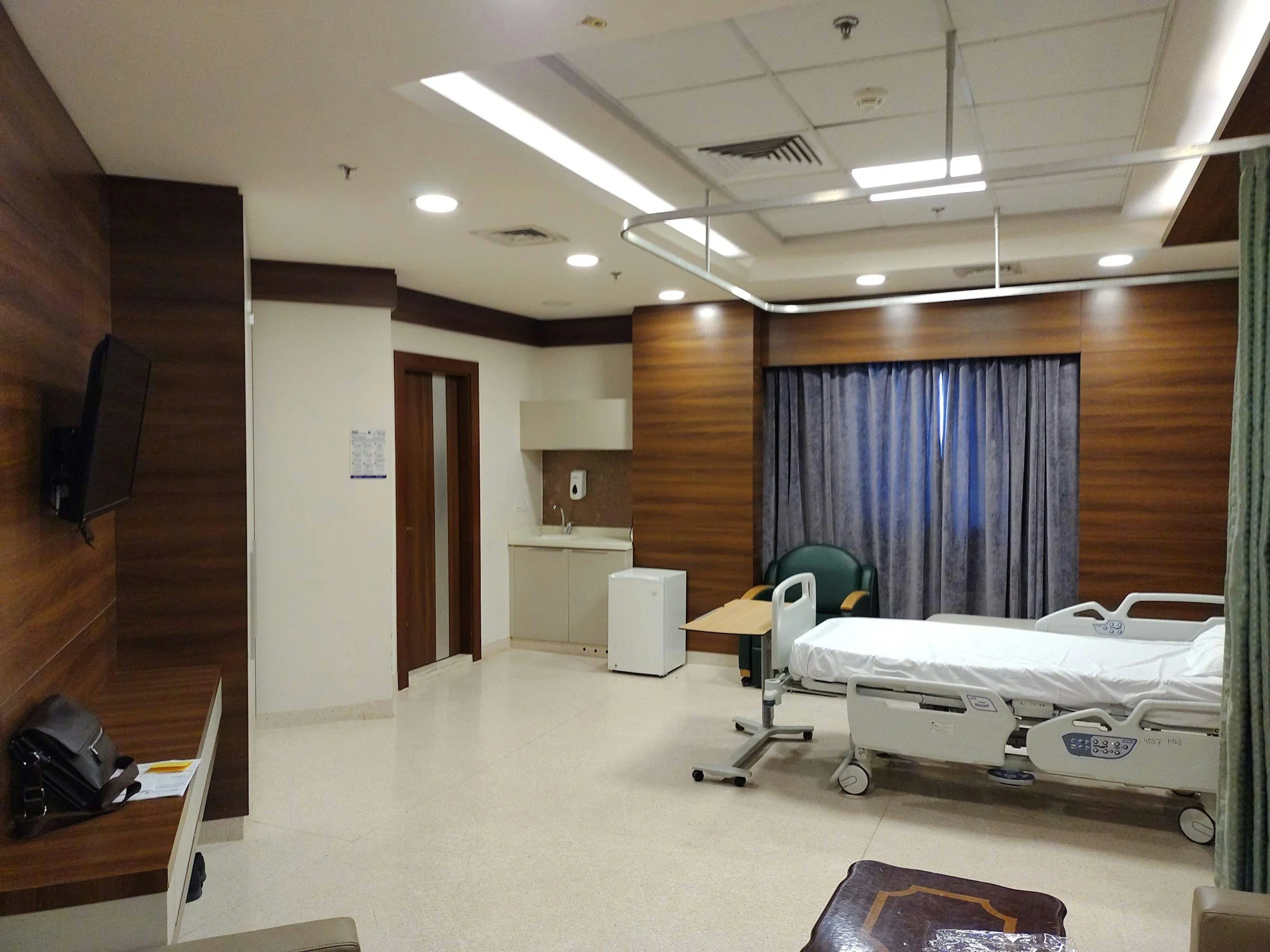 A hospital room with wood-paneled walls, a bed with white linens and a control panel at the foot, a green chair nearby, medical equipment, a sink, curtains, and a television mounted on the wall.
