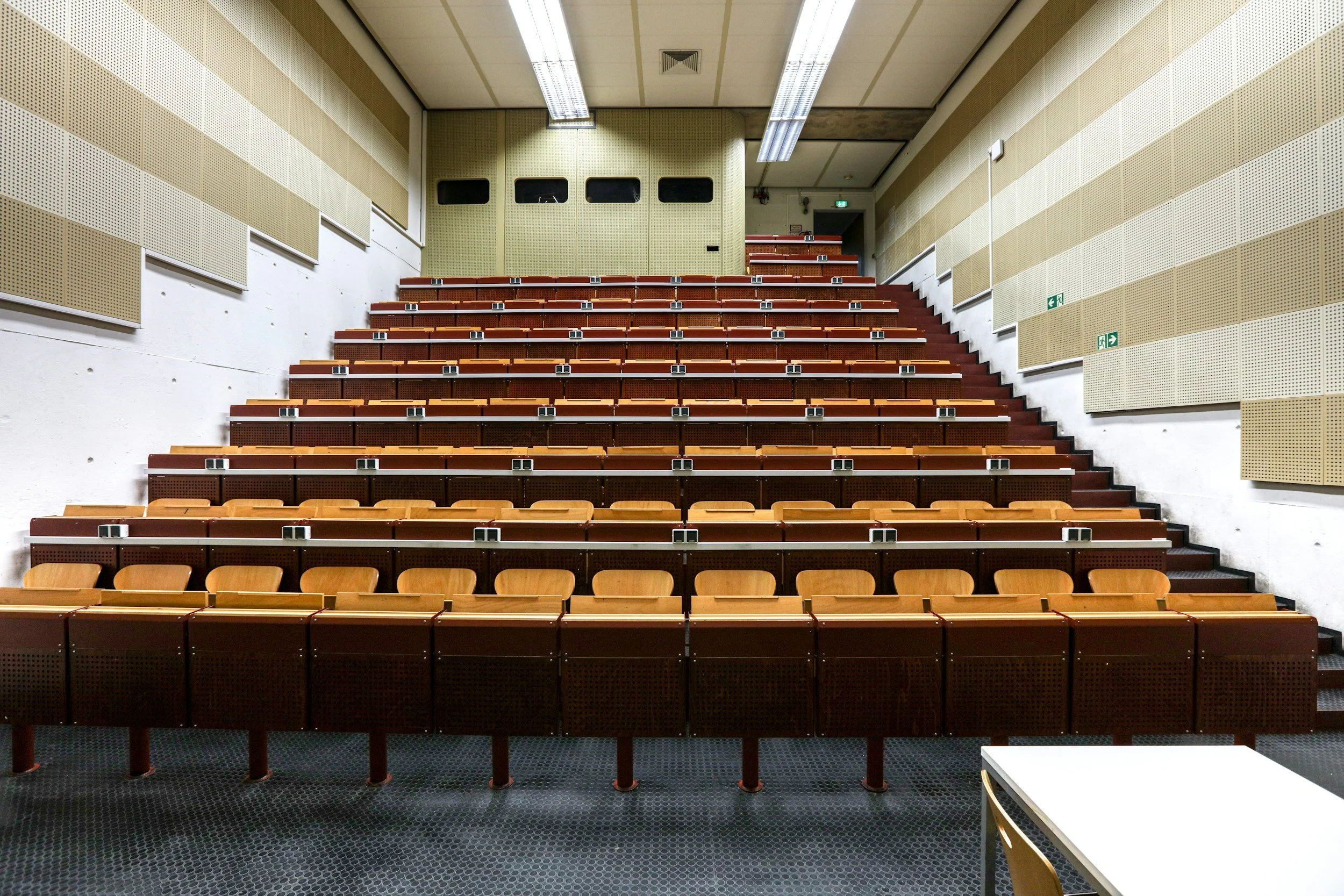 Empty auditorium with tiered wooden seating, beige acoustic panels on the walls, fluorescent ceiling lights, and two emergency exit signs.