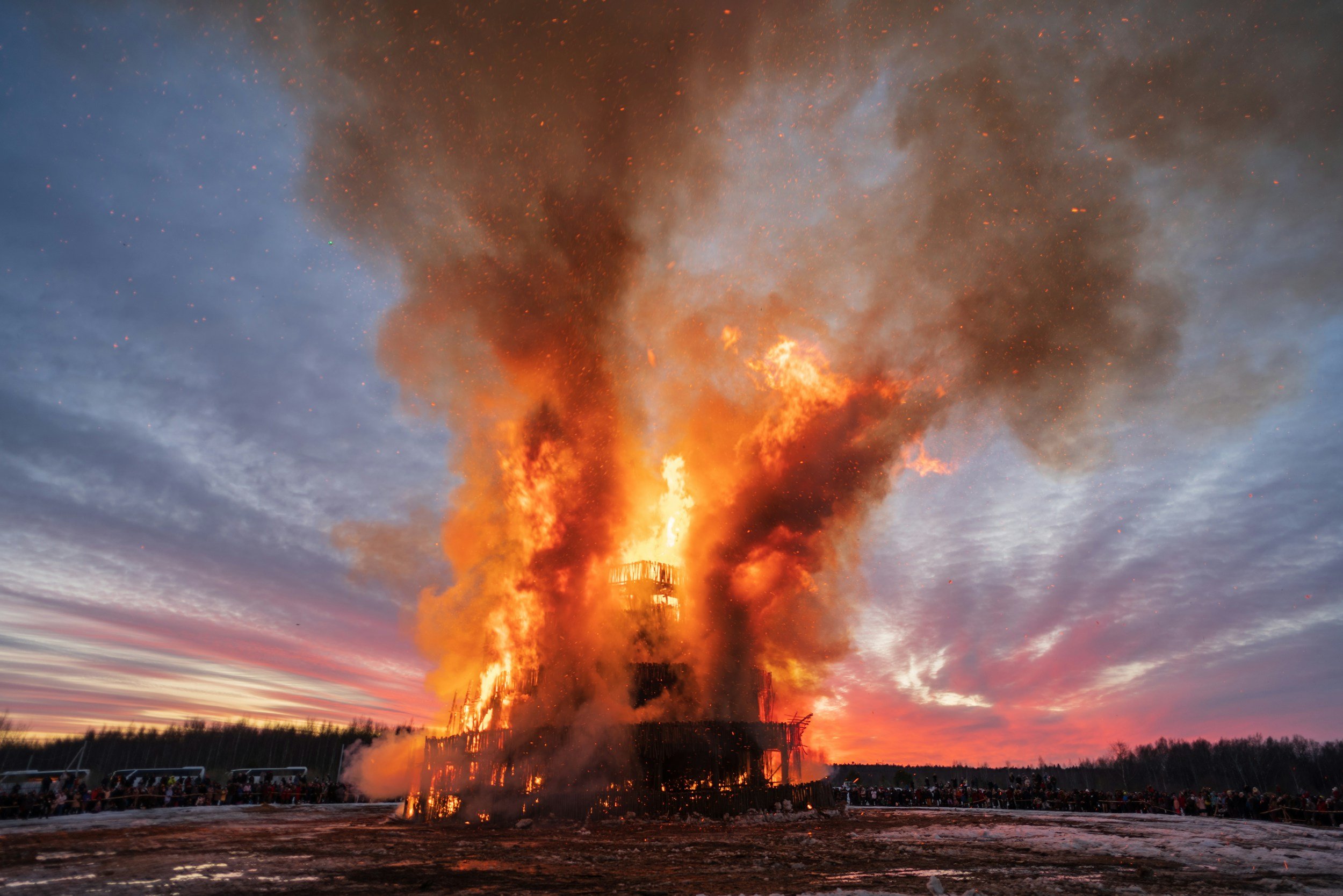 Fireworks or burning structure with intense flames and smoke against a sunset sky, with a large crowd of people and cars in the foreground.