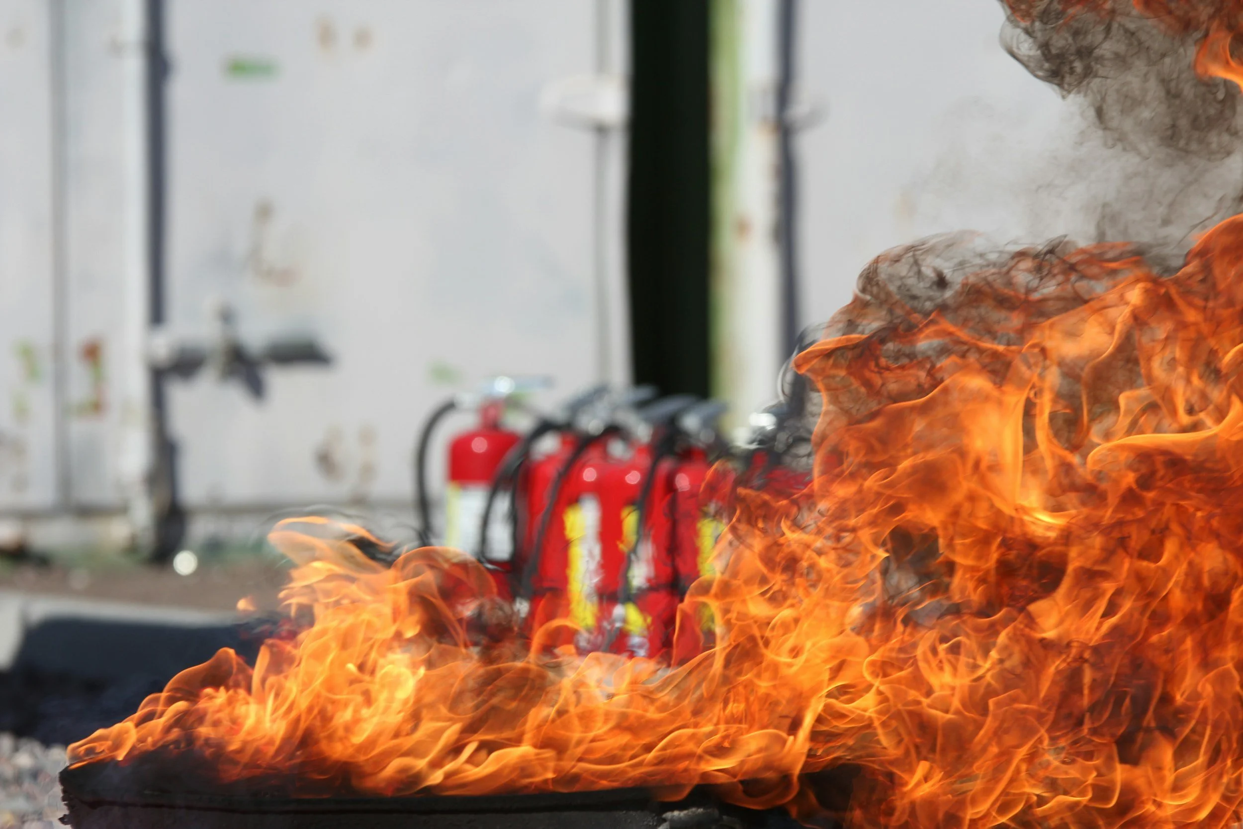 Flames and fire coming from a fire pit or controlled outdoor fire, with a background showing fire extinguishers and some laboratory or industrial equipment.