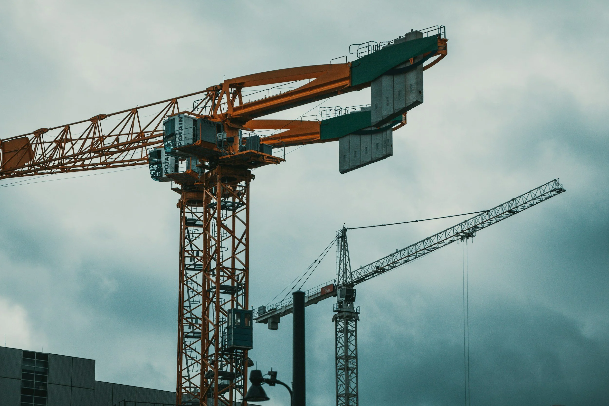 Construction cranes against a cloudy sky, with parts of a modern building at the bottom.