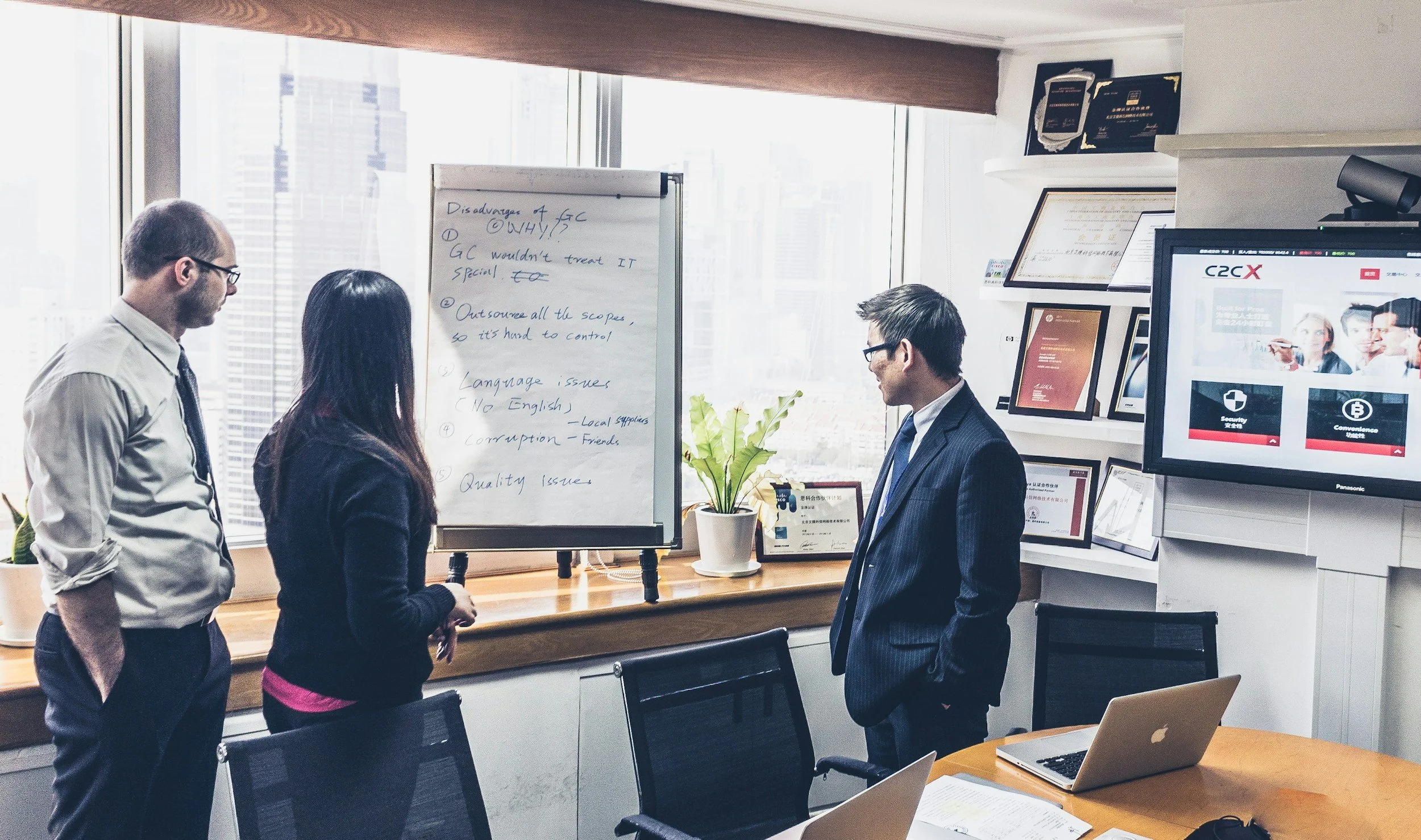 Four professionals engaged in a discussion in a modern office with large windows. One person is standing next to a whiteboard with notes about disadvantages of GC, outsourcing, language issues, corruption, and quality issues. There are multiple screens and framed awards or certificates on the wall.