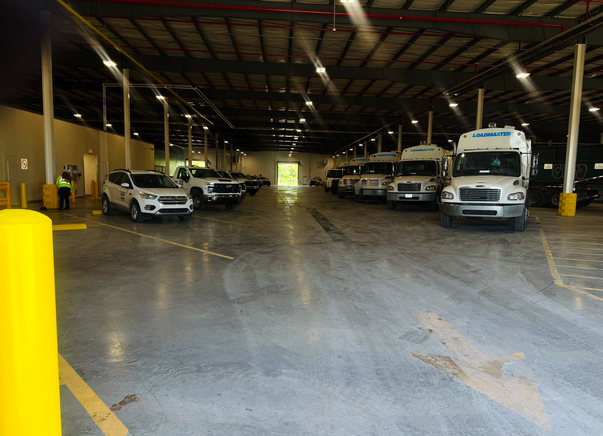 An indoor parking garage with a few cars and trucks parked along the sides, and a person wearing a safety vest standing near a wall on the left side.