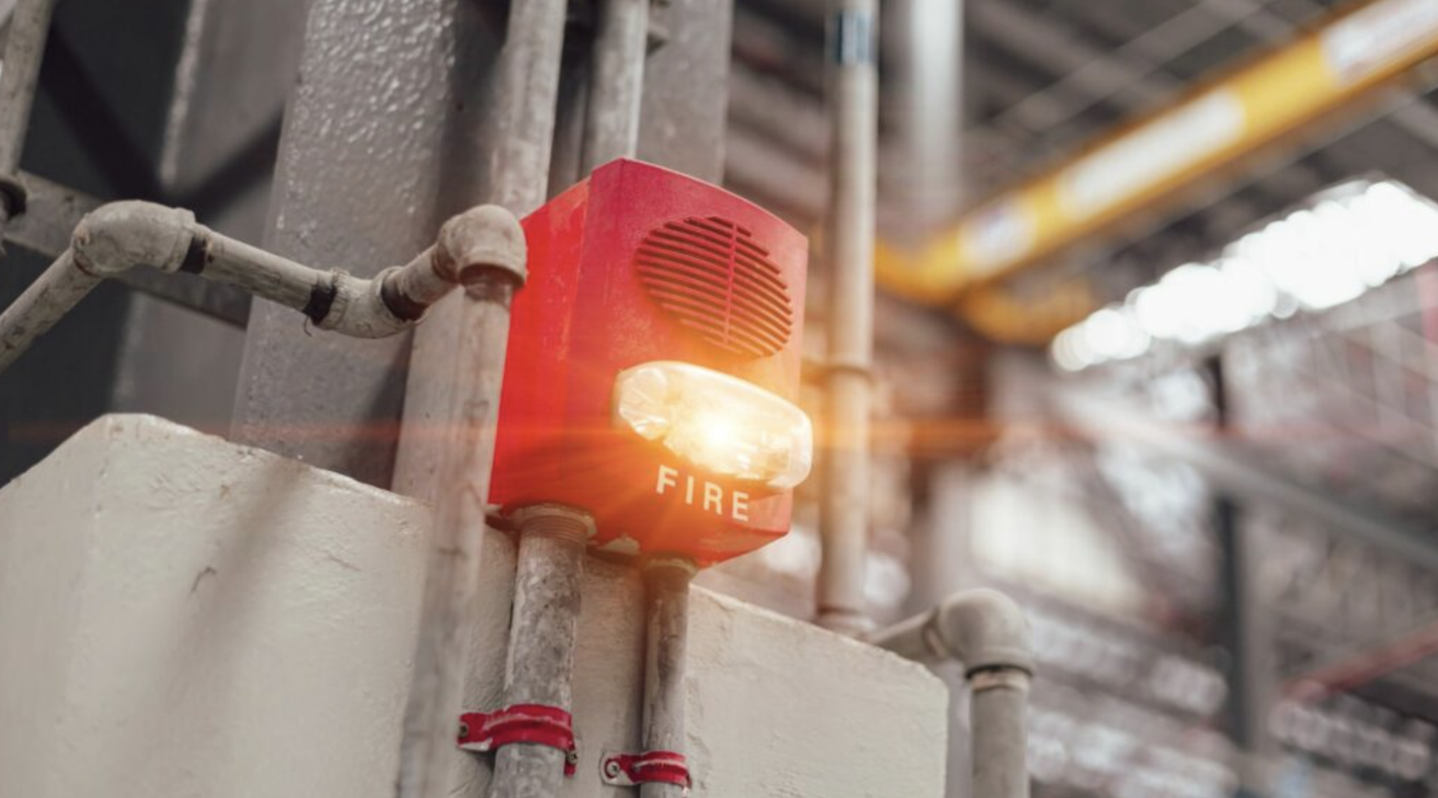 A red fire alarm with an orange indicator light and the word 'FIRE' on it, mounted on a white wall in an industrial or commercial setting with metal pipes and scaffolding in the background.