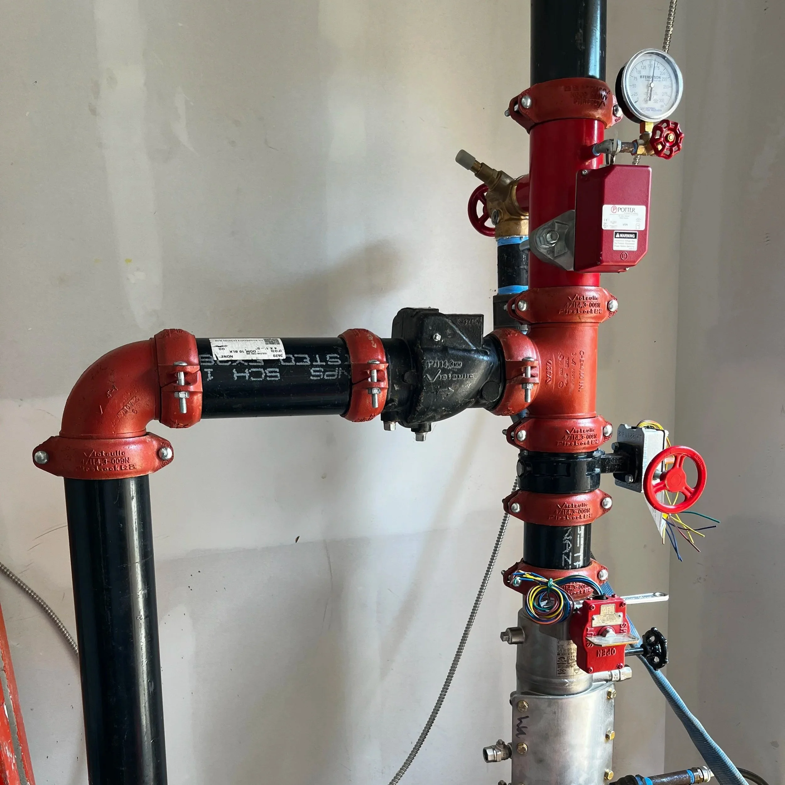 Indoor plumbing system with red pipes, a pressure gauge, and valves mounted on a white wall.