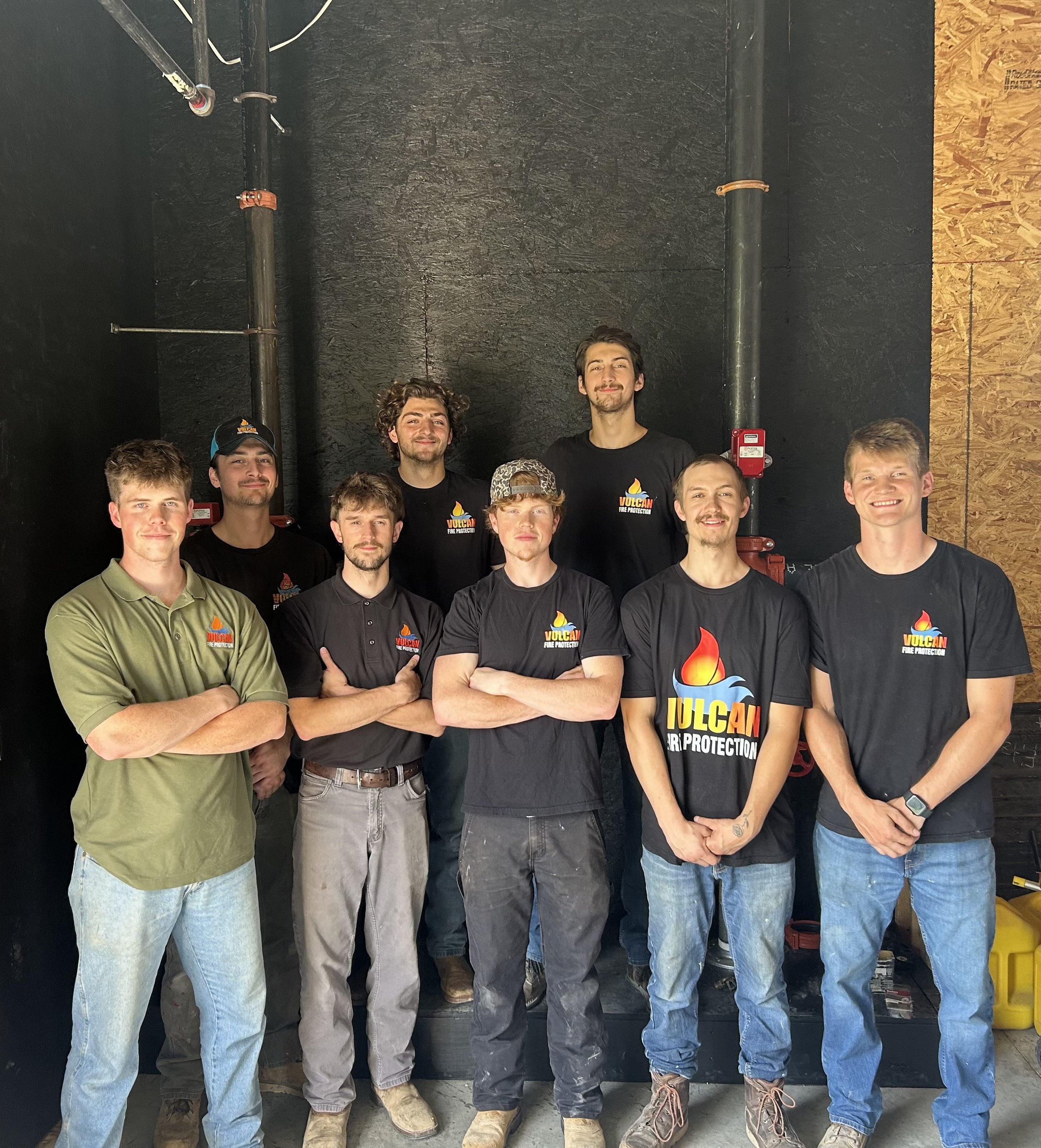 Group of young men standing together indoors, wearing black T-shirts with 'Vulcan Fire Protection' logo, smiling and looking at camera.