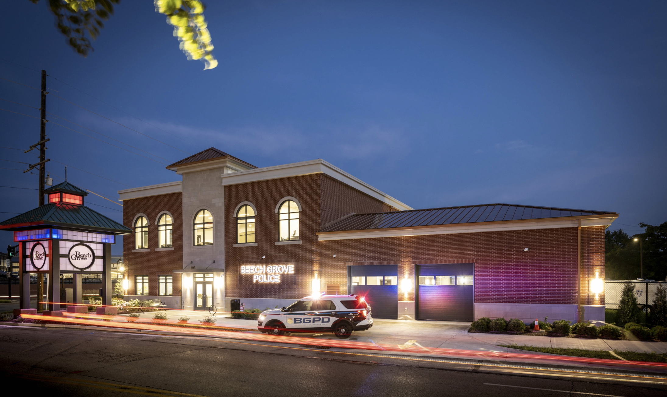 Nighttime street view of Beech Grove Police Department building with a police car in front, a lit sign, and a sky overhead with some clouds and blurred lights from passing vehicles.