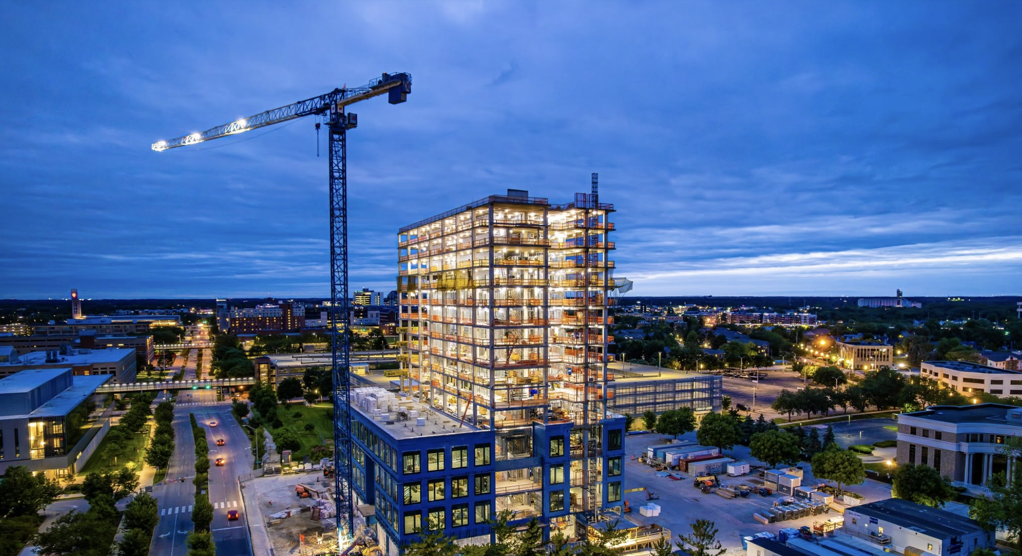 Night view of a multi-story building under construction with a tall crane beside it, city lights and clouds in the background.