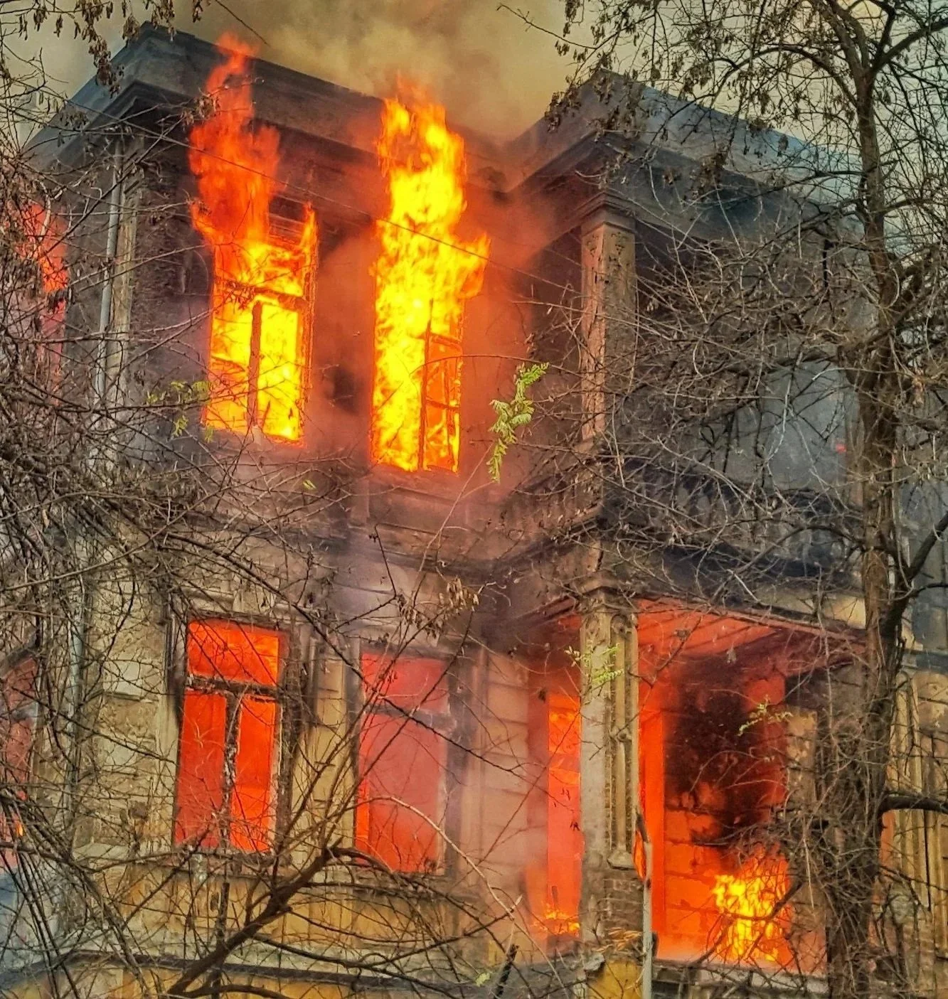 A two-story house is engulfed in flames, with fire coming out of the windows on the upper floor. Smoke is rising into the air, and the house is surrounded by leafless trees.
