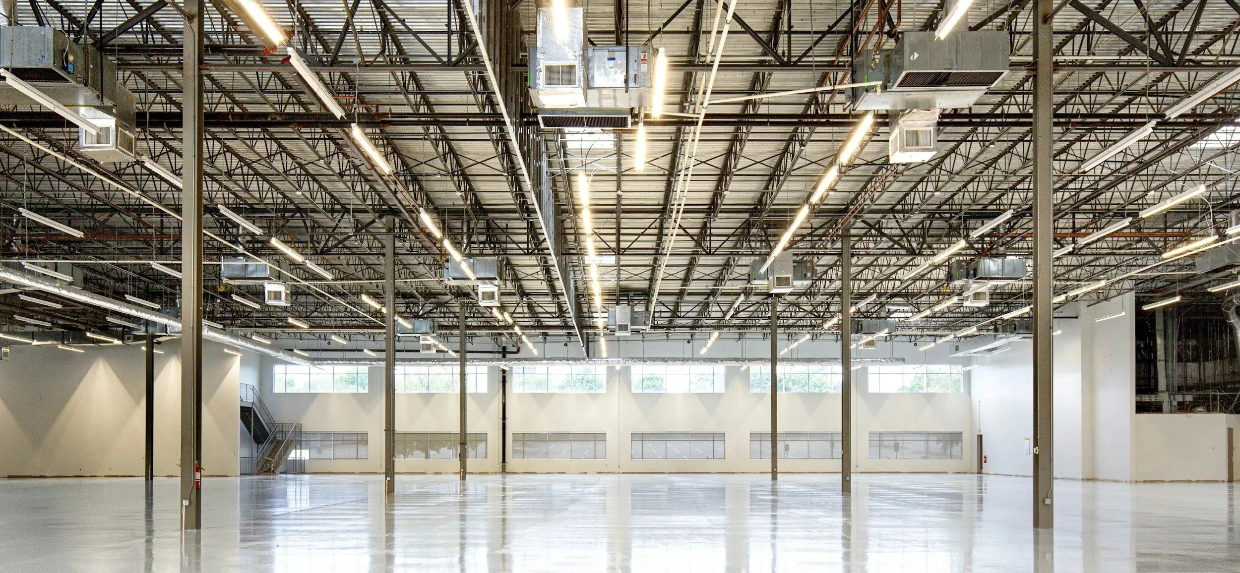 A large, empty industrial or commercial space with a high ceiling, exposed metal framework, industrial lighting, and large windows allowing natural light, with a polished concrete floor.