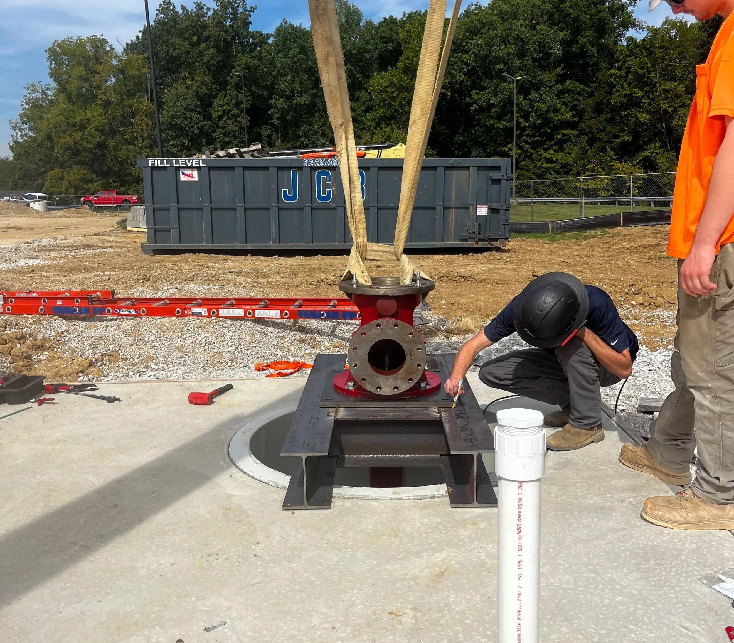 Construction site with a large gray dumpster labeled 'JCB' and a man in an orange vest working near a black pipe. Some trees and vehicles are visible in the background.