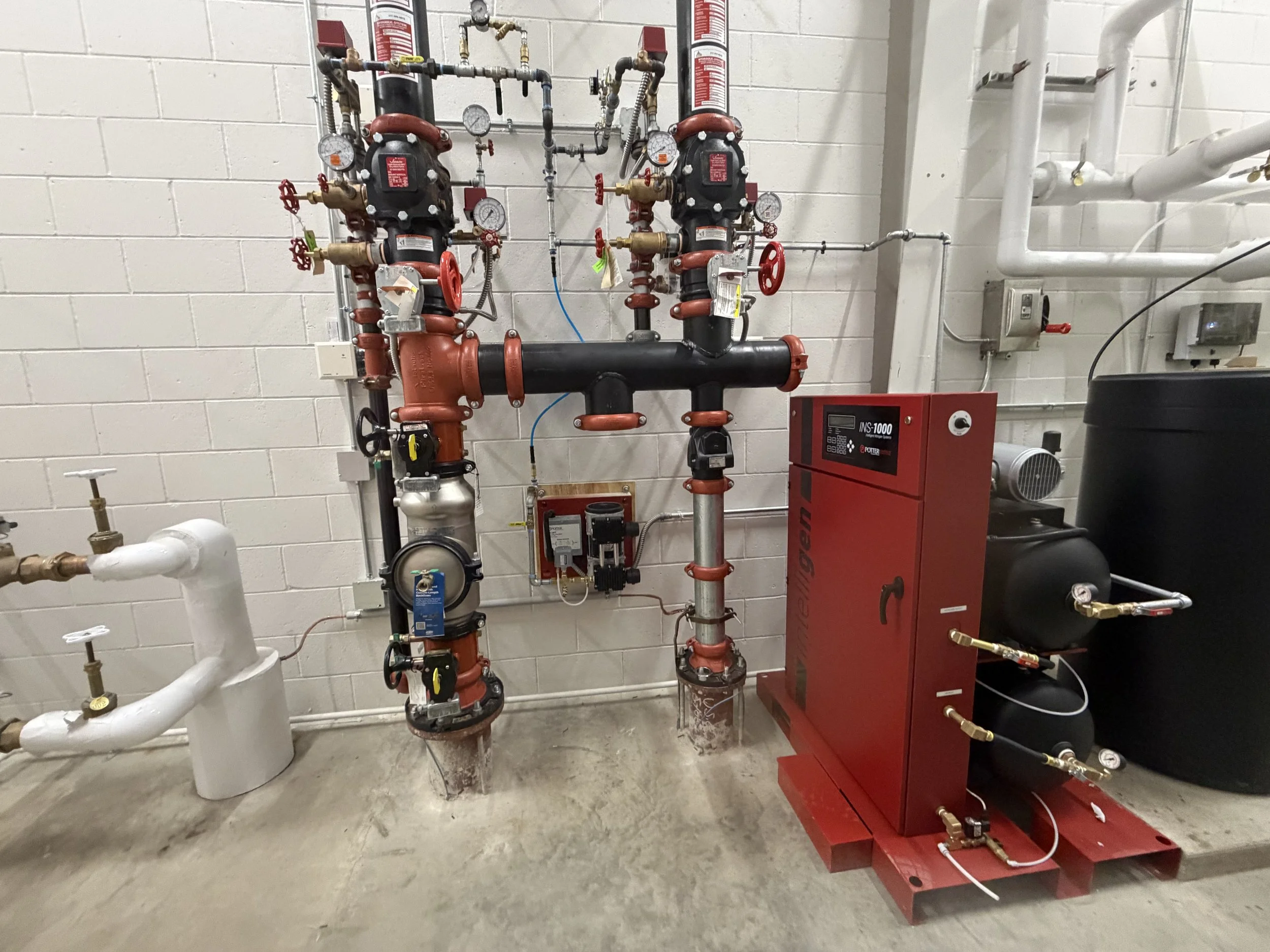 Industrial mechanical room featuring red and black pumps, pipes, gauges, valves, and control panels against a beige brick wall.