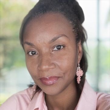 Close-up of a woman with braided hair, wearing a pink shirt and floral earrings, in front of a window with greenery outside.