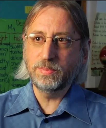 A man with shoulder-length gray hair, glasses, and a beard wearing a blue shirt, sitting in a room with a green poster and written notes in the background.