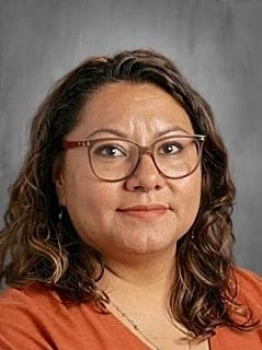 A woman with long dark hair, glasses, and earrings, wearing a green top, smiling slightly against a gray background.