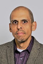 Photograph of a man with short hair and a goatee, wearing a gray blazer and a purple shirt, against a plain background.